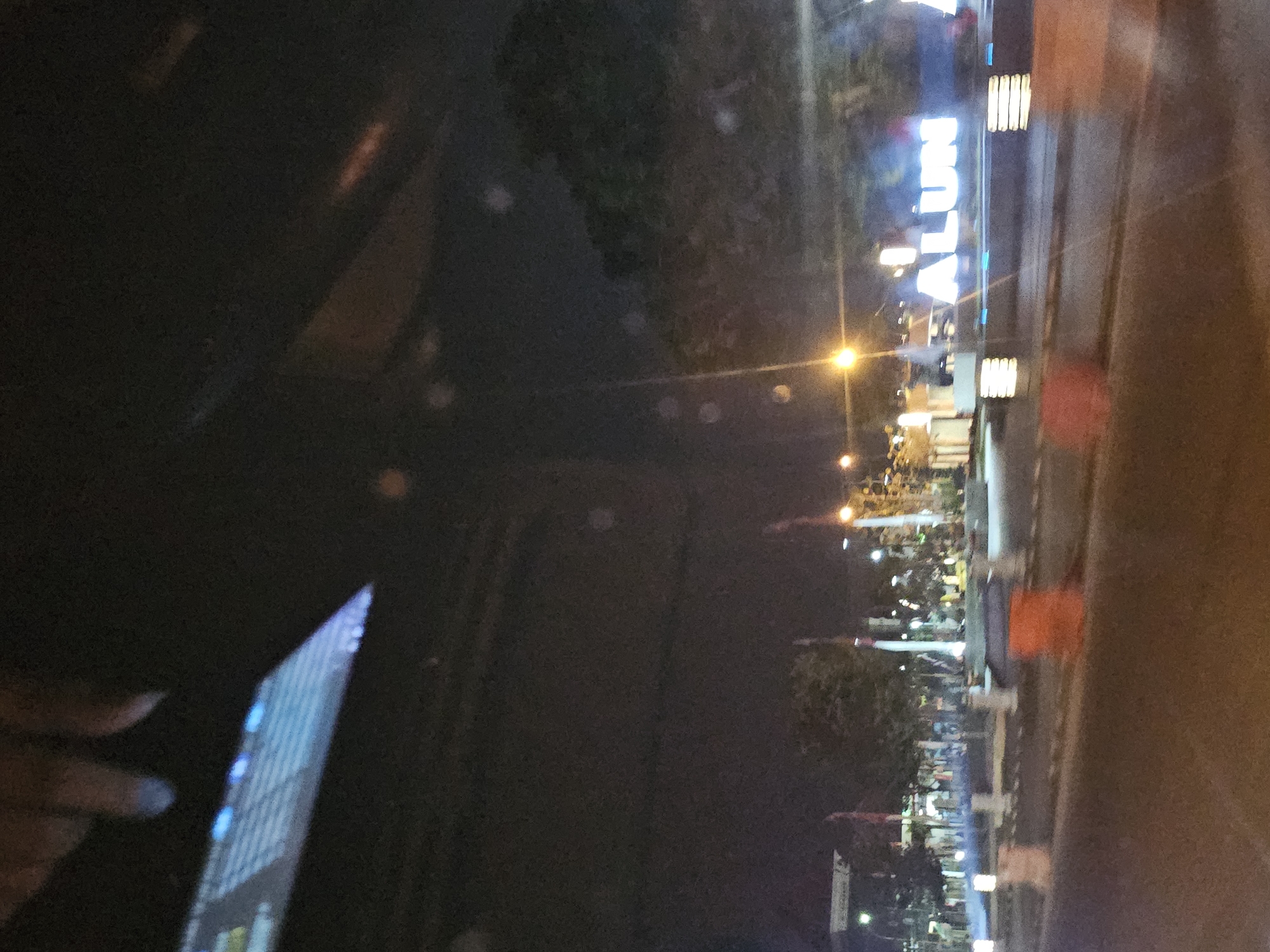 An out-of-focus nighttime shot captures a street scene in Tuban, Indonesia. In the foreground, a blurry road is visible, with orange traffic barriers suggesting a construction zone or event setup. Further back, illuminated by streetlights and possibly the glow from a digital sign, are elements of a public space. A prominent, large, illuminated sign spells out "ALUN-ALUN" which is Indonesian for "town square" or "city park." The "ALUN" portion is clearly legible, and the subsequent letters are partially visible and blurred. The background shows trees and buildings, also appearing indistinct due to the low light conditions and motion blur. The overall impression is of a busy public area at night, likely a town square in Tuban.