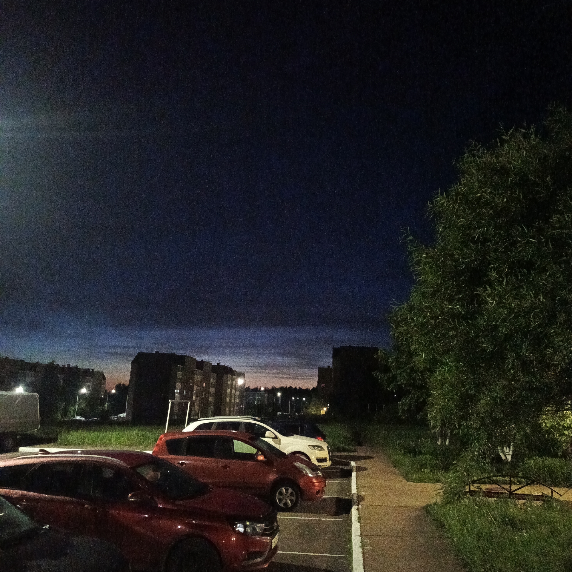 The image depicts a parking lot in Марковский, Russia, at night. Several cars are parked in marked spaces, including a red hatchback and a white SUV. Behind the cars are apartment buildings illuminated by streetlights. A large tree is visible on the right side of the frame. The sky is dark, with a faint glow near the horizon, suggesting a recent sunset or early sunrise. A sidewalk borders the parking spaces, with grass and foliage growing alongside it. The overall scene is quiet and typical of a residential area at night.