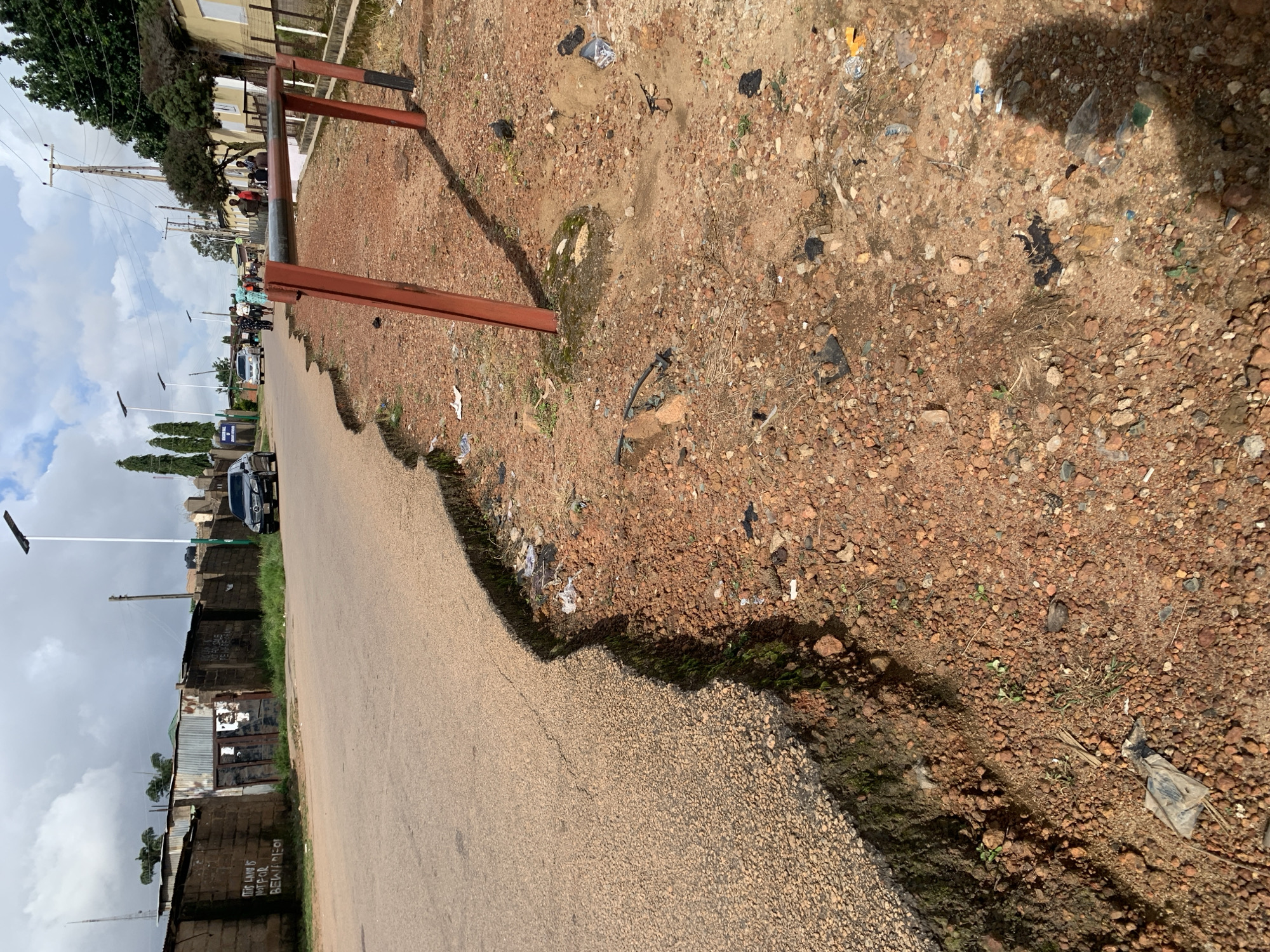 The image captures a street scene with a distinct erosion line along the edge of the asphalt road, exposing the reddish-brown soil beneath. The road appears to be a two-lane street with a worn surface. On the left side of the road, a low brick wall borders a property, with a corrugated metal gate and graffiti visible on the wall. Beyond the wall, structures with metal roofs are partially visible. A black sedan is parked on the right side of the street, further down the road, and several people are walking on the sidewalk and crossing the street in the distance. Streetlights are present along the road, and power lines are visible. The ground on the right side of the road is rough, covered with loose soil, small stones, and scattered debris, including plastic bags and wrappers. A red metal barrier or fence post is positioned on the right side of the road, with its base set in concrete. The sky is overcast with clouds. The scene suggests an urban or semi-urban environment.