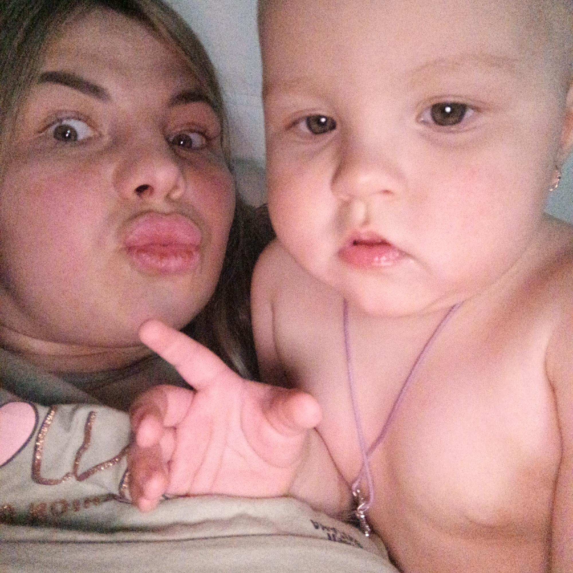 A selfie shot shows a fair-skinned adult and a fair-skinned infant in an indoor setting. The adult is on the left side of the frame, making a pouting face with wide eyes. The infant is positioned to the right, staring straight ahead. The infant is wearing a necklace with a pendant. The adult is wearing a T-shirt. The location is Unknown, Unknown.