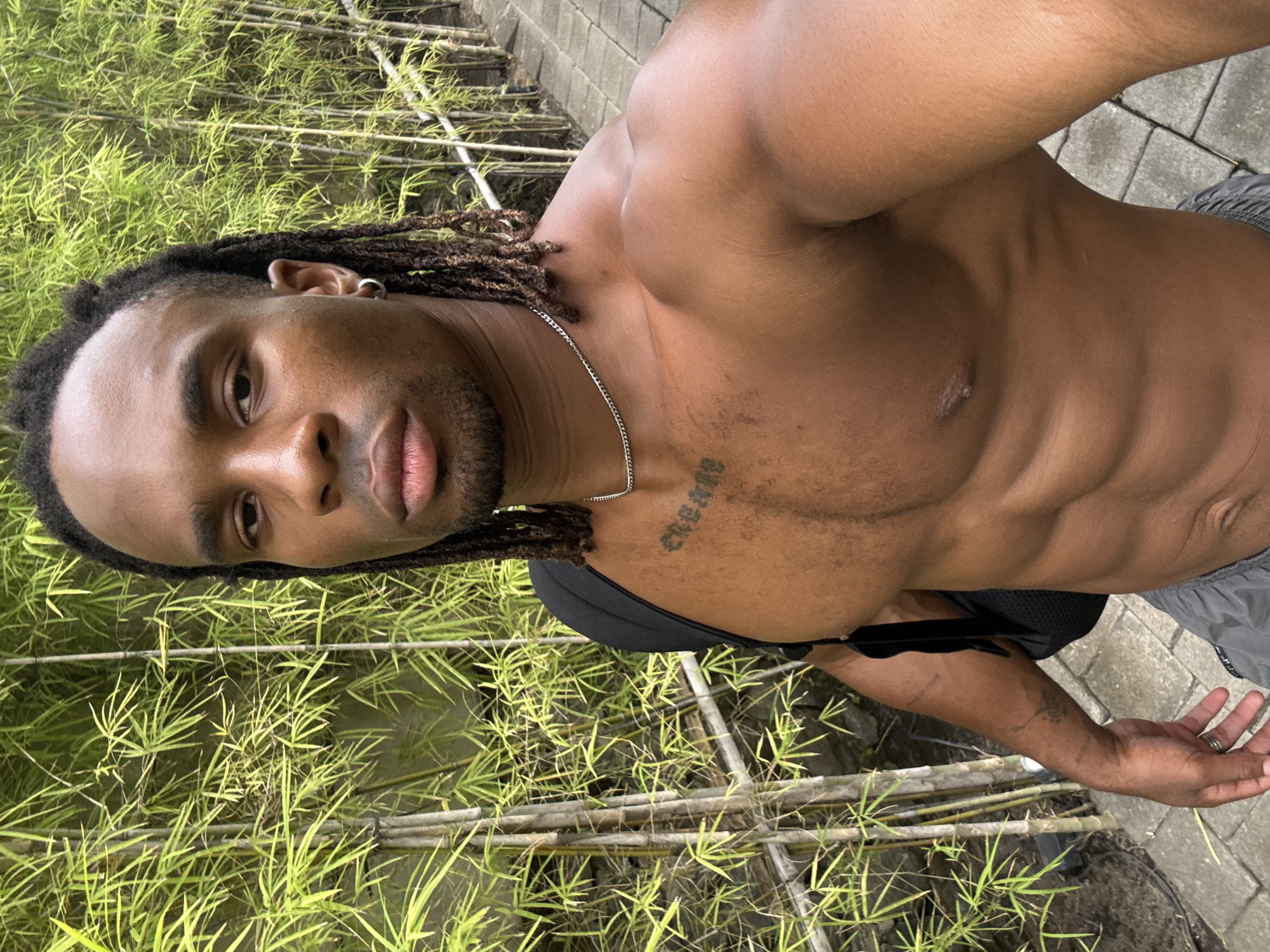 A shirtless man with dreadlocks stands outdoors, seemingly near a bamboo grove. He has a tattoo on his upper chest and is wearing a silver chain necklace. He is wearing a black top under the chain. The ground is paved with gray stone tiles. The man appears to be taking a selfie. The location is Unknown, Unknown.