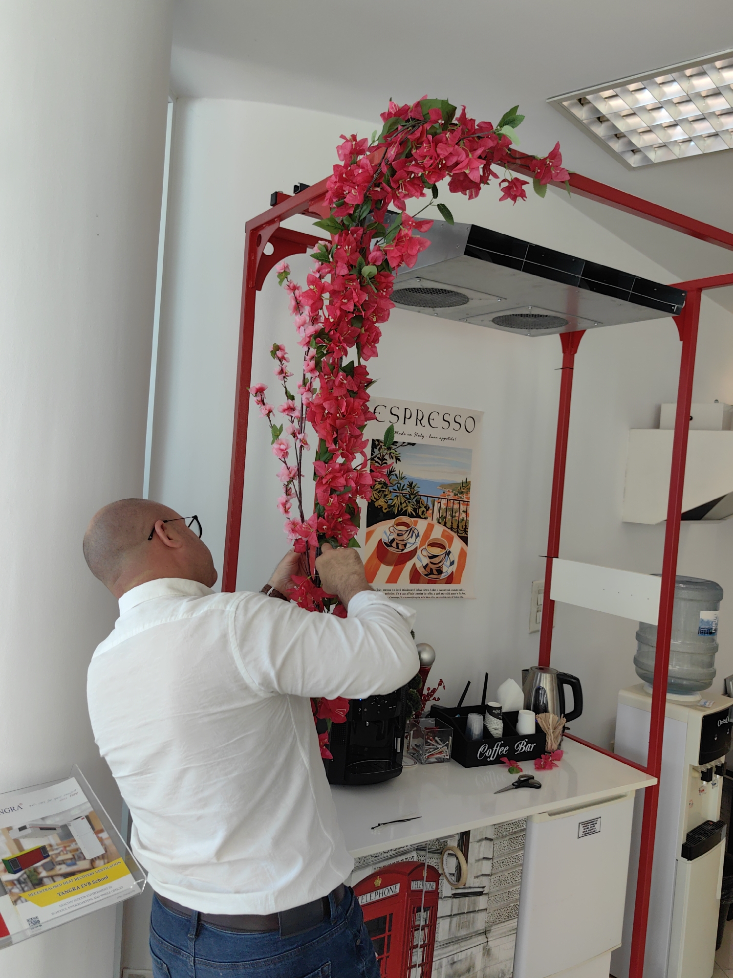 A man with a shaved head, wearing glasses, a white long-sleeved shirt, and blue jeans, is arranging red artificial flowers on a red metal structure. Behind him, there is a poster with the word "ESPRESSO" and an image of Italy. The environment includes a coffee bar setup with a kettle, coffee cups, and a water dispenser. A sign reads "Coffee Bar". A flyer with "TANGRA EVB School" visible is located to the lower left. The wall behind is white, and the setting appears to be indoors, possibly in an office or commercial space in Sofia, Bulgaria.