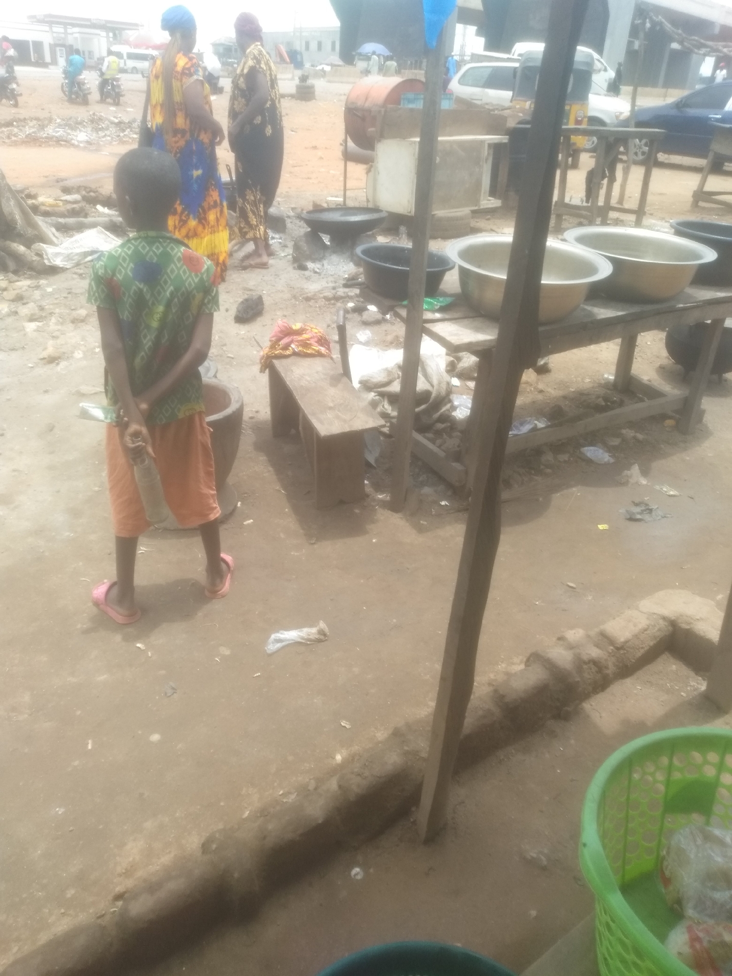 In an outdoor scene of what appears to be a marketplace, a young child stands with their back to the camera, wearing a green patterned short-sleeved shirt and orange shorts, with pink flip-flops on their feet. The child is holding a clear plastic bottle. To the left of the child, there is a large, hollowed-out cylindrical object that resembles a mortar. Further back, two women are visible; one wears a colorful yellow and blue patterned dress and a blue headscarf, while the other is in a black and gold patterned dress with a dark headscarf. They are standing and conversing. In the background, several motorcycles are parked, and a white van and other vehicles are present, suggesting a public area. The ground is a mix of dirt and debris. In the mid-ground, a wooden table holds several large metal bowls, some appearing empty and others possibly containing items. A smaller wooden bench is situated behind the child. A green plastic basket containing items wrapped in plastic is visible in the foreground on the right. The overall environment is weathered and appears to be a bustling, possibly informal, commercial setting.