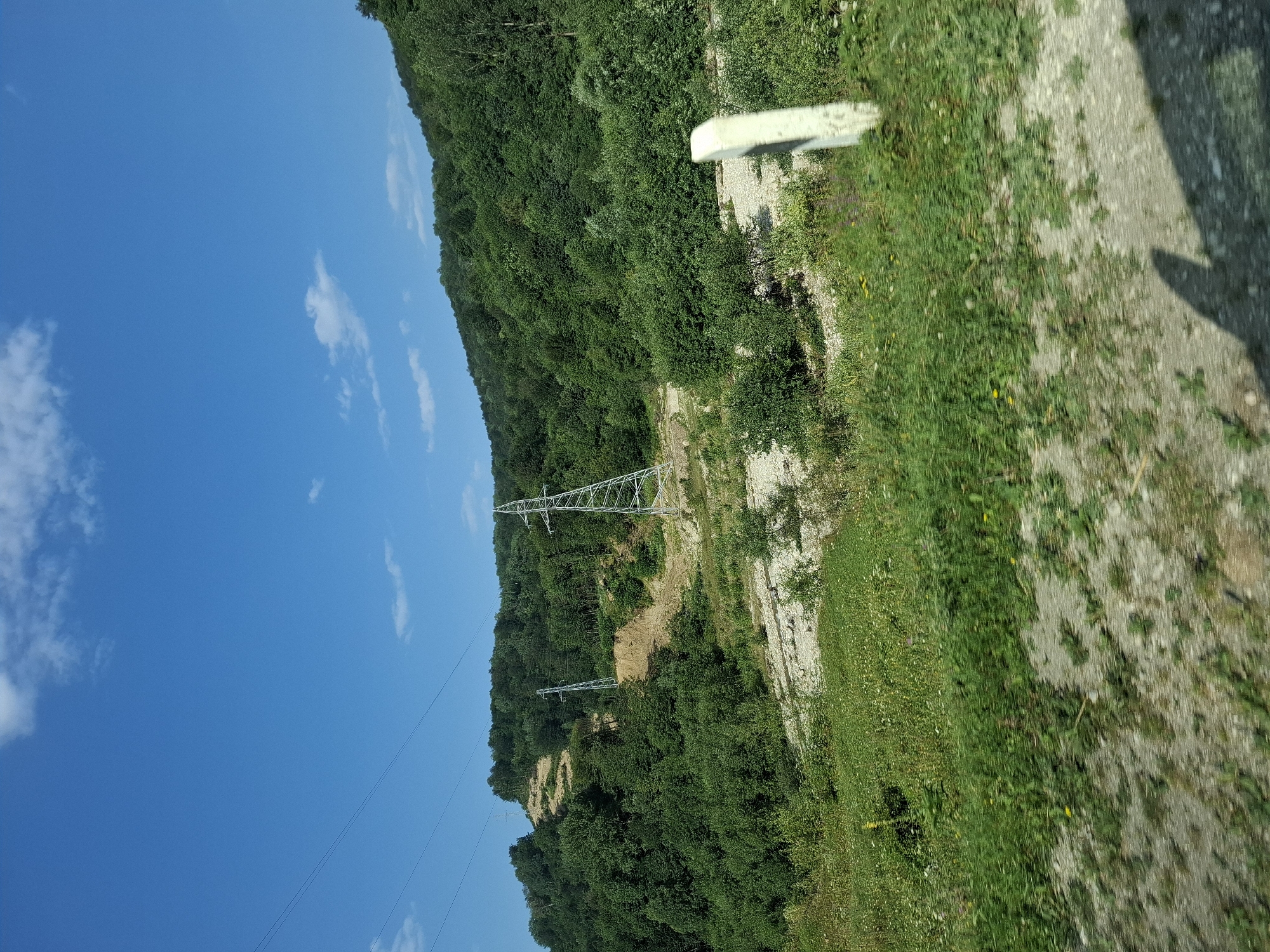 The image depicts a hilly landscape with dense green vegetation. Power transmission towers are visible on the slopes, indicating the presence of electrical infrastructure. A partially cleared area on one of the hills suggests recent construction or maintenance work. The foreground shows a gravelly ground with sparse green grass and some small yellow flowers. A white marker post stands to the right of the frame. The sky is blue with a few scattered white clouds. The overall scene suggests a rural or semi-rural environment, possibly near a road given the blurred foreground and the presence of the marker post. The image is not specifically located in the city of Dyshne-Vedeno, Russia, nor does it contain any identifiable people or actions.