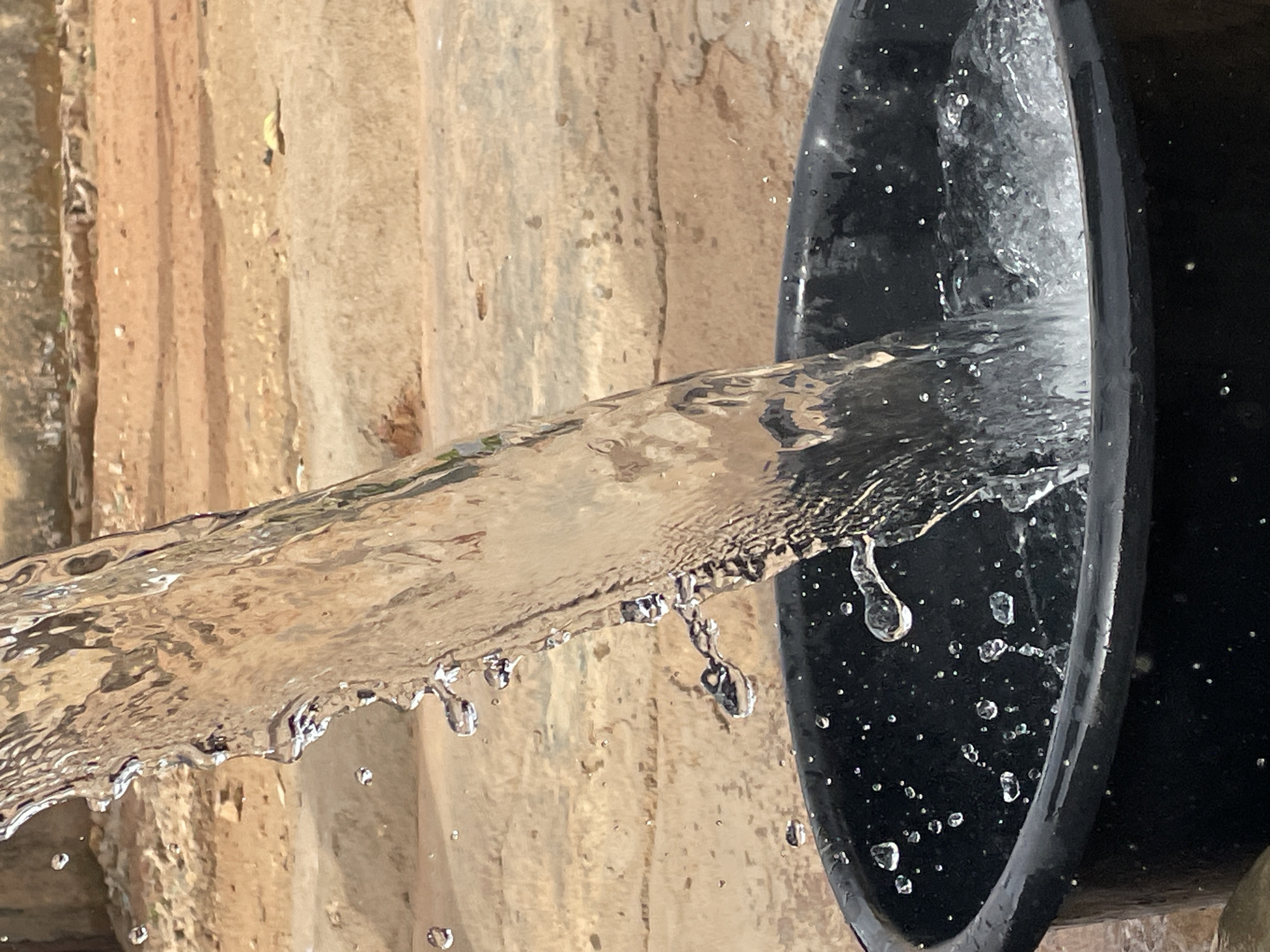 A stream of water is pouring from an unseen source into a black container. The water appears relatively clear, with droplets detaching from the main flow. The container is round and catches the stream. The background reveals a textured wall, composed of vertical panels with a rough, earthy tone. There is no evidence to substantiate the location as Jos, Nigeria. The scene lacks any identifiable people.