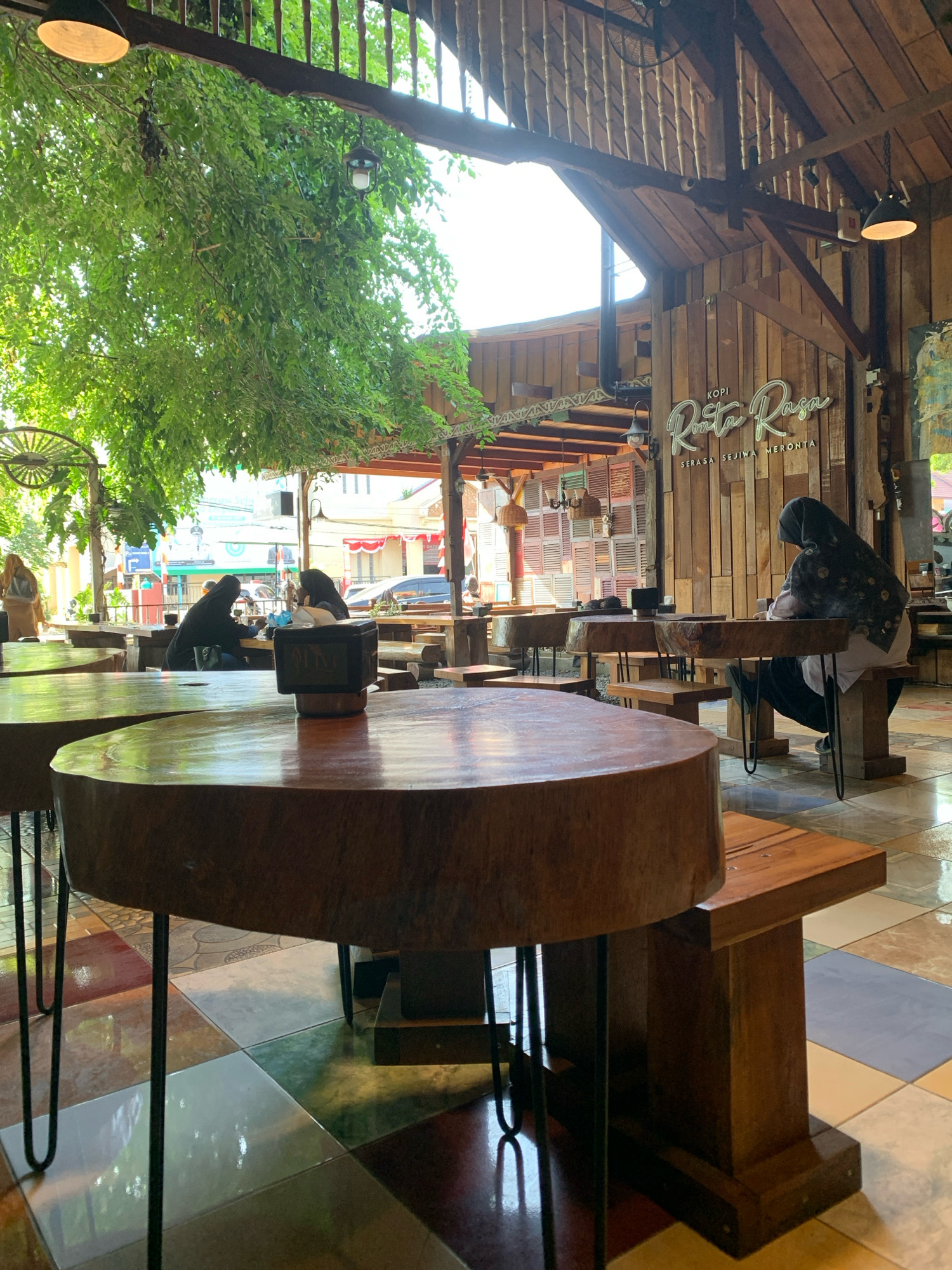 The image depicts the interior of a cafe with a rustic, wooden design. Large, round wooden tables with metal legs are prominent in the foreground. In the mid-ground, several people are seated at tables. Two individuals wearing hijabs are visible at one table, engaged in conversation. Another person wearing a hijab is seated alone at a table in the background, facing away from the camera. The cafe features exposed wooden beams, wooden paneled walls, and hanging lamps. A sign on the wall reads "Kopi Rinta Rasa". Green foliage from an outdoor tree drapes into the cafe through an open-air section. The floor is tiled with a mosaic pattern of various colors and textures. The scene appears to be a casual gathering or dining setting. The context provided indicates the location is in the city of Lhokseumawe, Indonesia.