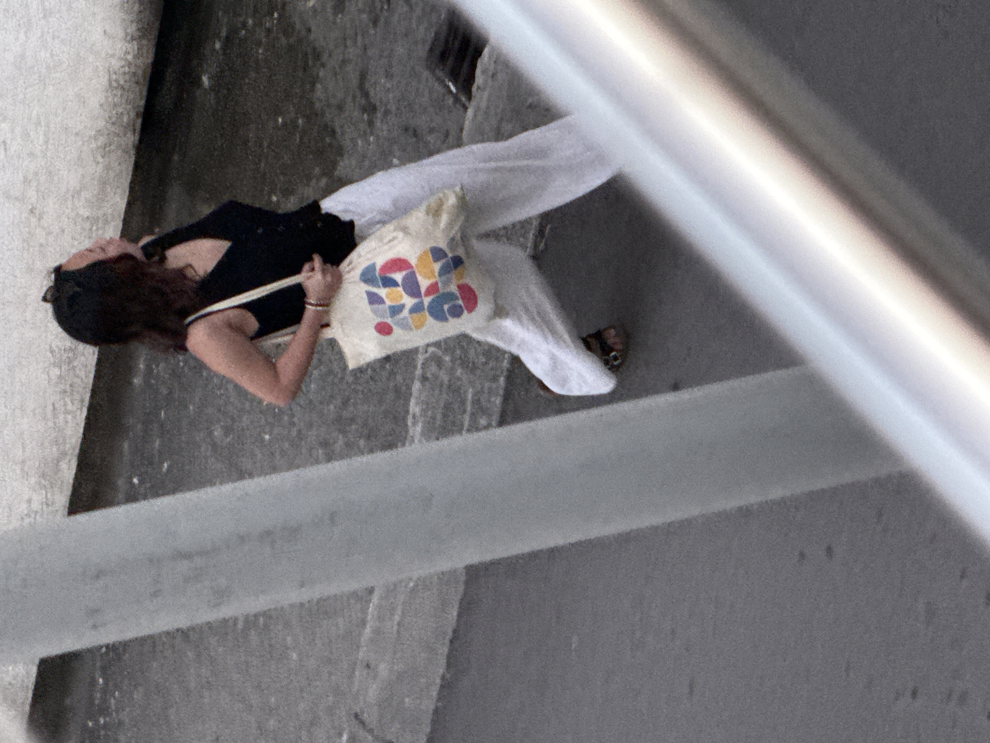 A woman with dark, shoulder-length hair, wearing a black v-neck sleeveless top and loose white pants, walks away from the camera on a paved surface. She is carrying a light-colored tote bag with a colorful geometric pattern across her left shoulder. The bag contains a dark object, possibly a wallet or phone. Her right arm is bent and her hand rests on the bag strap. She wears sandals. A pale, textured wall is behind her, and a tall, grey cylindrical pole is to her left, partially obscuring her. The ground appears wet in patches. The scene is observed from an elevated angle. The location is identified as Budva, Montenegro, suggesting an outdoor urban environment.