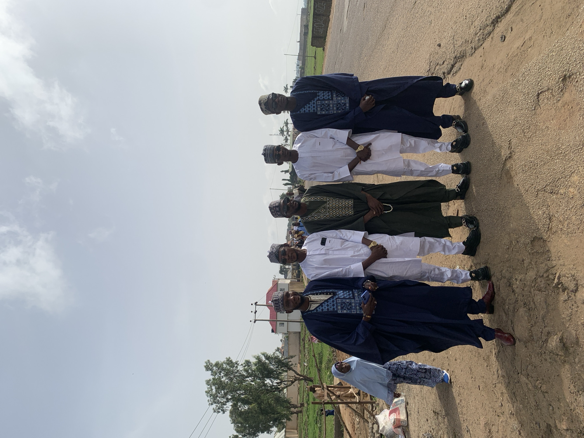 Five men stand in a line on a paved surface in Naraguta Hausawa, Nigeria. The men are all wearing traditional Nigerian attire. Two men wear navy blue robes with intricate designs, two wear white robes, and one wears a green robe with a similar design. The men wear matching caps and dark shoes. Behind the men, a few buildings are visible, along with a utility pole. A tree is also visible. In front of the men, a woman in a light blue dress and patterned shawl stands partially obscured. Some trash, possibly building materials, and a wooden structure are also visible in the foreground. The sky is overcast.