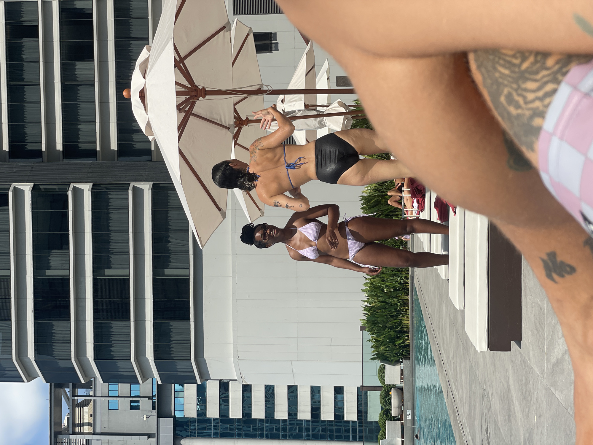 The image depicts a scene at a rooftop swimming pool in Bangkok, Thailand. Two women in swimwear are standing near the pool. One woman is wearing a black swimsuit bottom and a blue bikini top, and has tattoos on her back and arm. The other woman wears a white and lavender checkered bikini. An individual with a tattoo is partially visible in the foreground. In the background, there is a modern high-rise building with a distinctive facade and white umbrellas. The pool area is surrounded by foliage and lounge chairs.