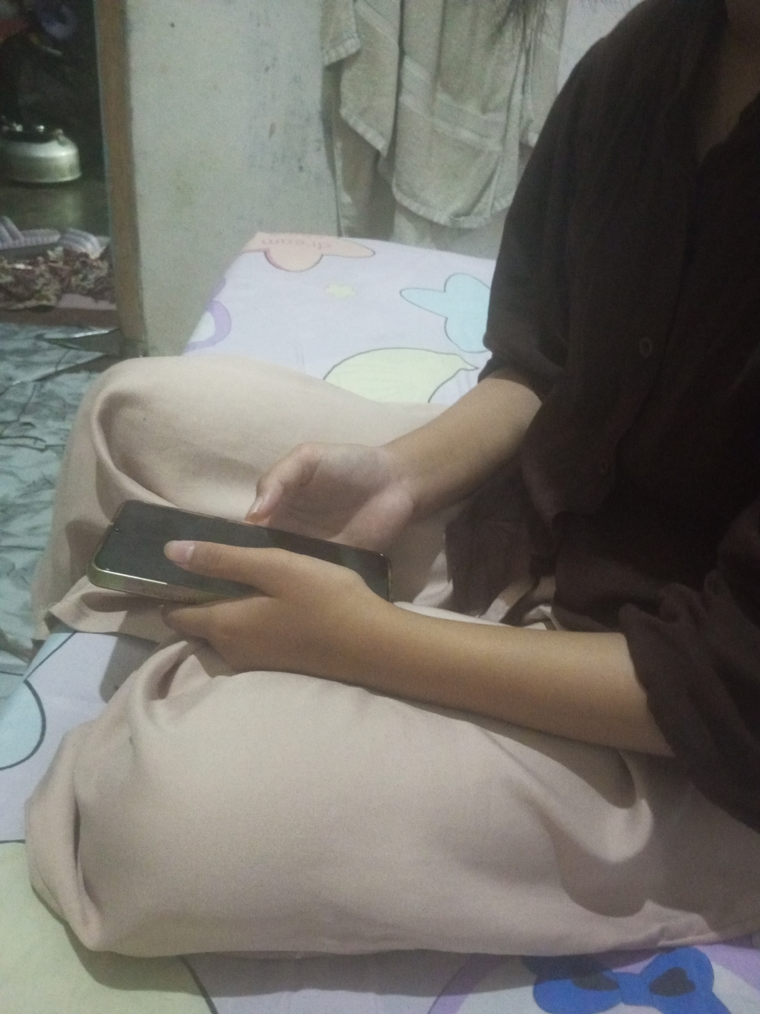 The image shows a person sitting cross-legged on a bed in what appears to be a residential interior in Andongsili, Indonesia. The person is wearing a dark brown long-sleeved shirt and light beige pants. Their hands are resting on a smartphone, which is held horizontally. The bedsheets have a light color with a pattern of cartoonish pastel-colored flowers. In the background, there is a portion of a light-colored wall and some fabric hanging from it. To the left of the frame, partially visible, is a corner of what looks like a wooden cabinet with a silver kettle on top, suggesting a domestic setting, possibly a bedroom or a living area within a home in Andongsili.