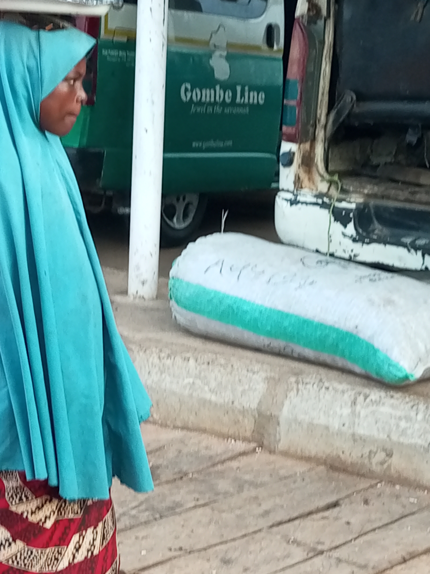 A medium shot captures a woman wearing a turquoise hijab and a red and gold patterned skirt, standing outdoors near a green bus with the text "Gombe Line" visible. To the right of the woman and the bus, a white sack with a green stripe lies on the ground. A white pillar is positioned between the woman and the bus. The scene appears to be an outdoor public space, possibly a transportation hub or market area within Gombe, Nigeria. The lighting suggests it is daytime, with natural light illuminating the subjects.