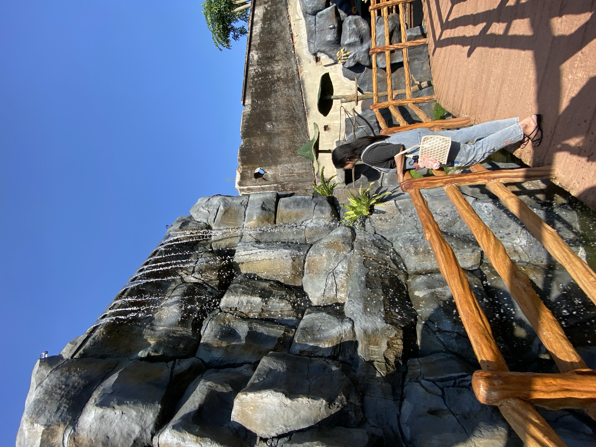 A woman is seated on a wooden railing near a rocky waterfall structure under a clear blue sky. The location is likely in Mangli, Indonesia. The woman has long dark hair and is wearing jeans and a dark top. She is holding a small purse with pink accents. The rocky waterfall structure is grey and wet, with water trickling down its surface. The wooden railing appears to be part of a bridge or walkway. A partially visible building with weathered exterior is located behind the woman and the waterfall structure.