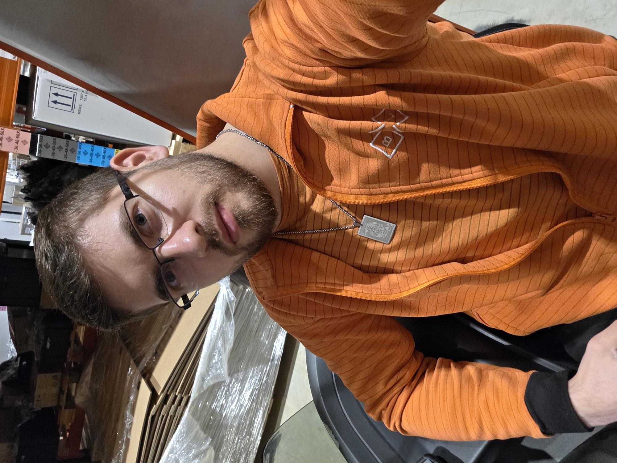 A man with short brown hair, a beard, and glasses is wearing an orange striped hooded jacket and a silver necklace with a rectangular pendant. He is sitting in a black chair. Behind him, there are stacks of wrapped cardboard boxes and shelves with boxes labeled with sizes. The background is slightly out of focus. There is no specific evidence to place the image in Plovdiv, Bulgaria.