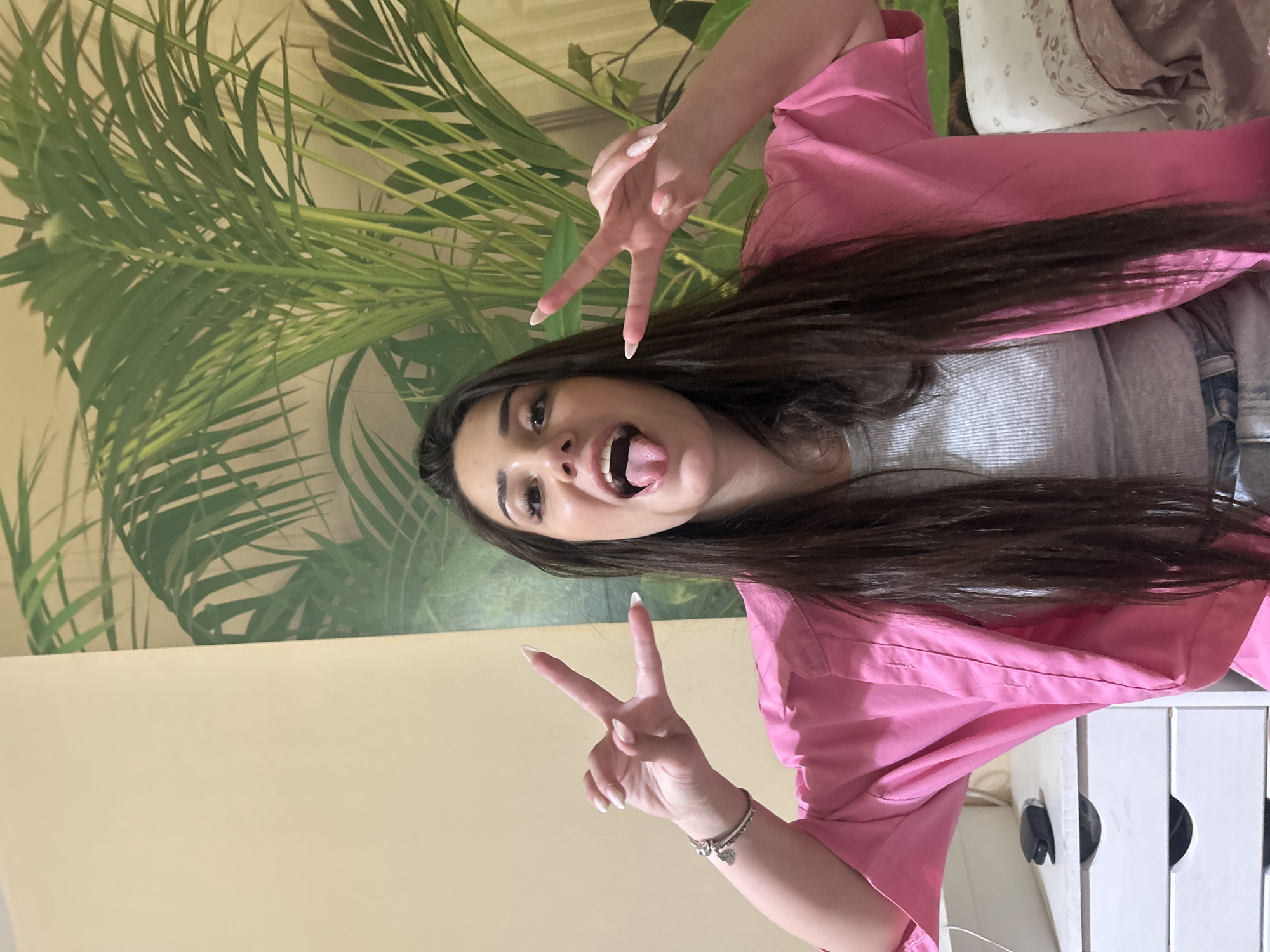 The image displays a young adult female positioned centrally, directly facing the viewer. She has long, straight dark brown hair and a light complexion. Her facial expression is playful, with her tongue extended. Both of her hands are raised, each forming a "peace" sign with the index and middle fingers extended. Her fingernails are long, almond-shaped, and painted with a light or natural color, potentially a French tip style. She is wearing a light grey ribbed crop top layered under an open, vibrant pink collared shirt. A small visible portion of light-wash denim jeans is present at the bottom of the frame. On her left wrist, she wears two bracelets, one of which is a charm bracelet with multiple charms, including a heart shape.
The background is divided into two sections. The left side features a plain, light beige wall. The right side exhibits a wall covering or wallpaper depicting large, green tropical plant fronds, resembling a palm. In the lower left foreground, a white dresser or nightstand with two visible drawers is present. On its surface, a white cable and a black car key fob are visible. A small section of patterned fabric or bedding in muted tones is visible in the lower right foreground. The specific geographical location, including the city of Plovdiv, Bulgaria, cannot be determined from the visual information provided in the image.