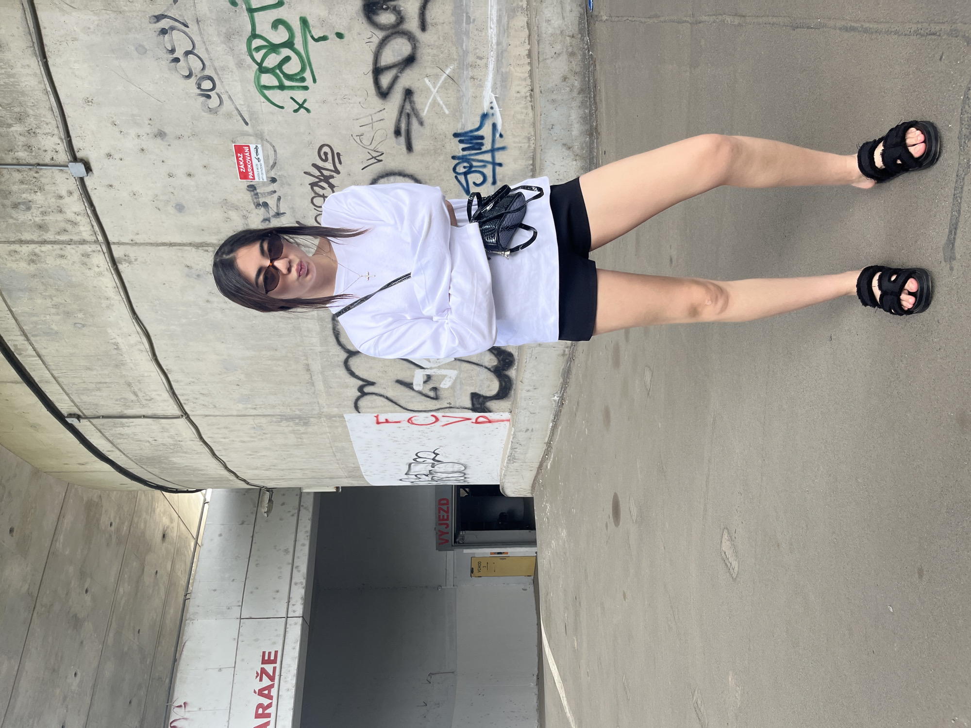 A woman stands with her arms crossed in front of a graffiti-covered concrete wall. She is wearing a white long-sleeved shirt, black shorts, black sandals, and sunglasses. A small black crossbody bag is slung across her chest. The background shows a concrete structure, possibly part of a parking garage, with a sign that reads "VÝJEZD" (Exit) in red letters above a yellow door. The location is the parking garage of the Olympia shopping center in Plzeň, Czechia.
