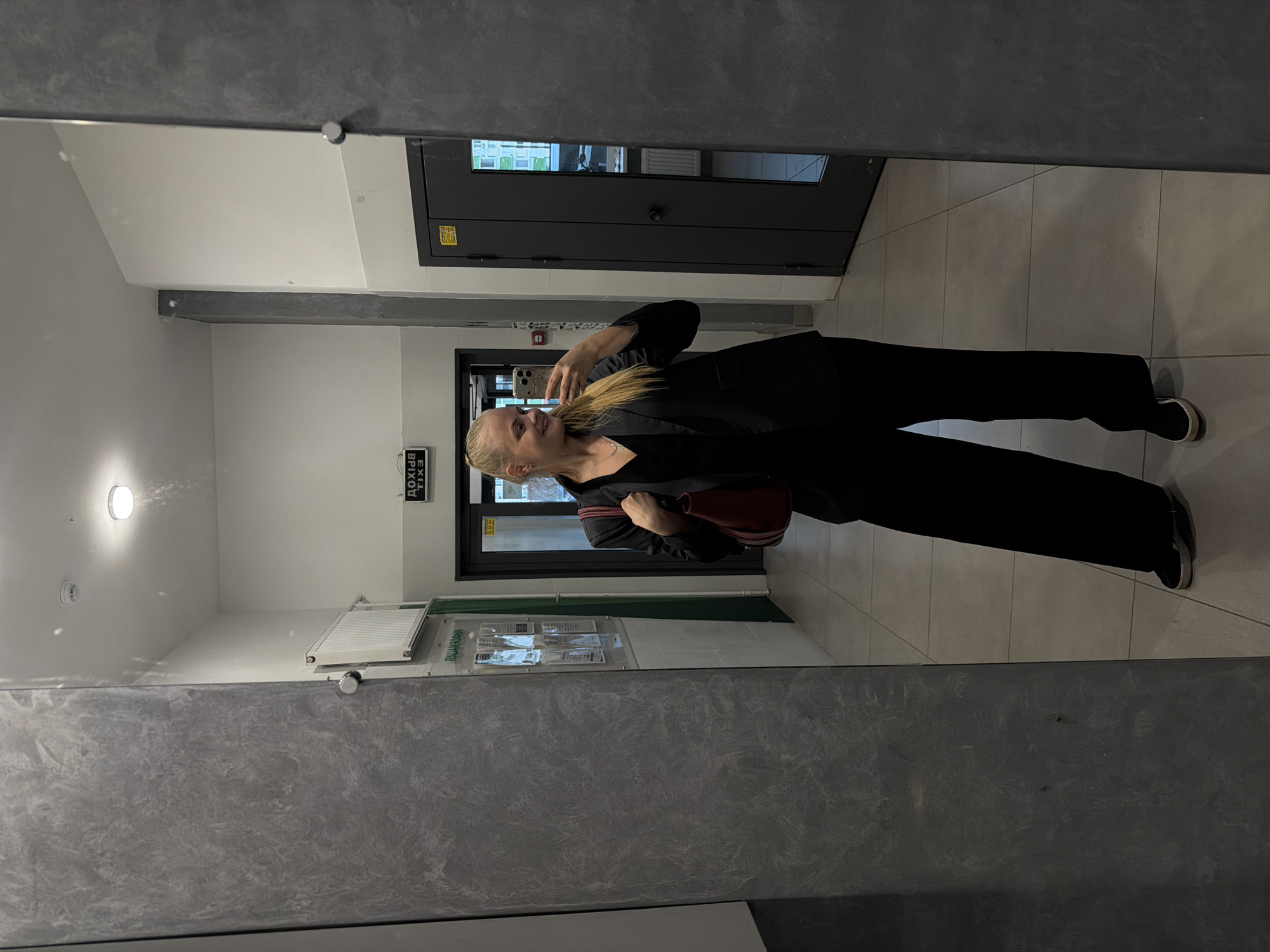 A blonde woman in a black blazer and wide-leg black pants takes a mirror selfie in a modern, well-lit hallway. She is holding a dark red handbag and a smartphone. The hallway has light grey tiled floors and white walls. On the left wall, there is a large gray textured pillar and a radiator. On the right wall, there is a dark gray door with a window panel. Above the door on the right, a sign in Russian reads "ВЫХОД" (Exit). The overall scene suggests an indoor common area, potentially in a residential building or office space in Izhevsk, Russia.
