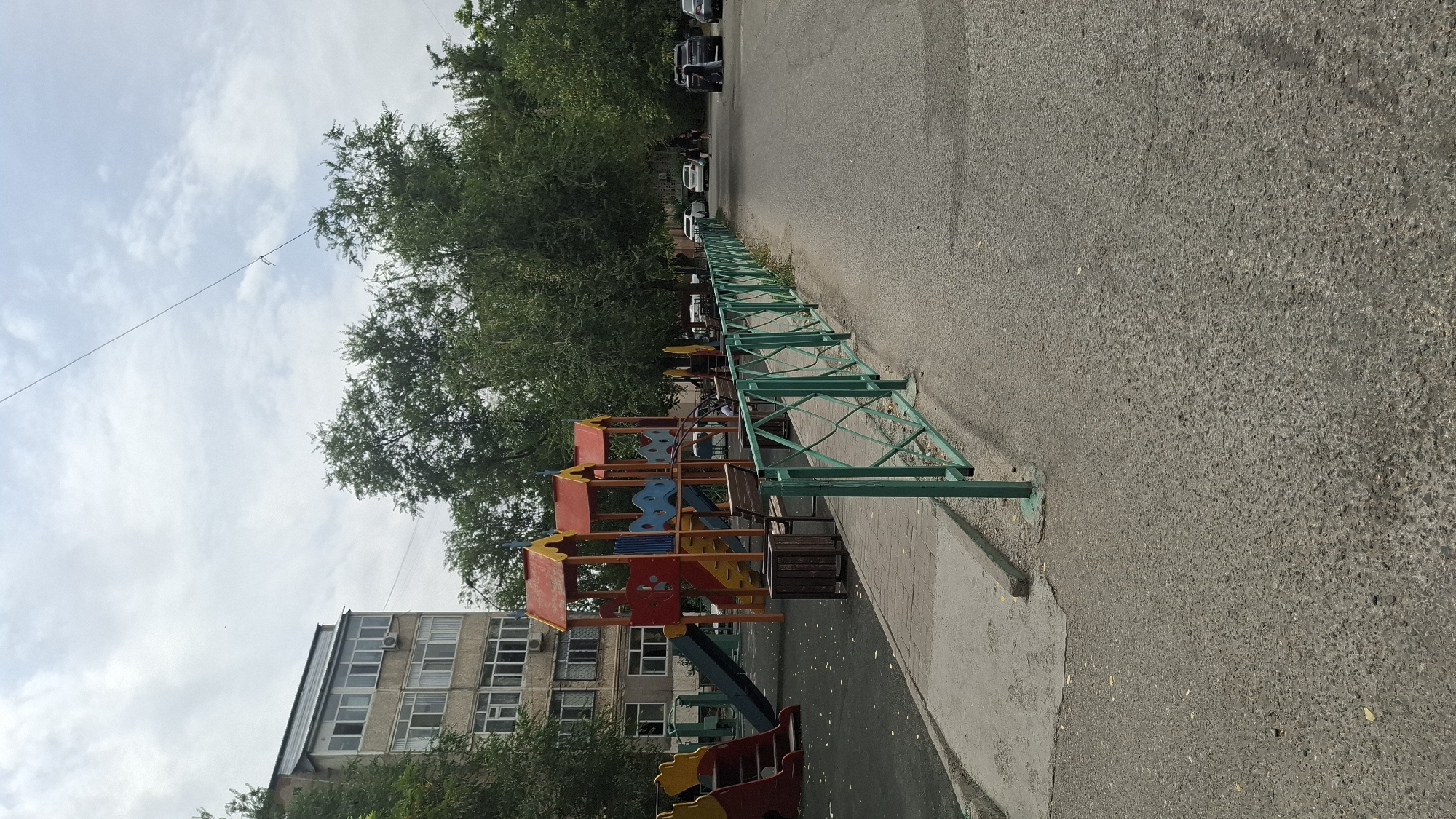 The image depicts a playground and a street scene in an urban environment, likely in Shymkent, Kazakhstan. A multi-colored playground structure with slides and climbing elements is visible on the left side of the image, set against a backdrop of a multi-story apartment building. Trees with dense foliage line the area between the playground and the street. A paved walkway leads from the playground towards a green metal fence that separates the playground area from an asphalt street. Several cars are parked along the street, and a few individuals are walking in the distance. The sky is overcast with scattered clouds.