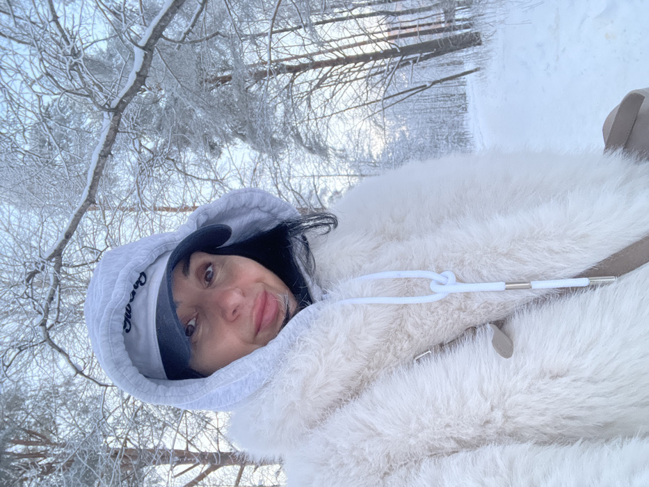 The image shows an adult female subject positioned outdoors in a snow-covered environment within Syasstroy, Russia. She is depicted from the chest up, looking directly at the camera with a neutral expression and a slight smile. She has dark hair and is wearing a light grey hooded sweatshirt with the hood up, underneath which she wears a dark baseball cap with light-colored lettering on the front. Over this, she wears a cream-colored, long-pile faux-fur coat or jacket. A white drawstring, featuring a decorative knot and two silver-colored cylindrical aglets, hangs centrally on her front. A light-colored strap, consistent with a bag, is partially visible over her right shoulder. The background consists of a dense winter forest with numerous trees heavily laden with white snow and frost. A snow-covered path or track is visible receding into the right side of the frame. The sky is overcast and bright.