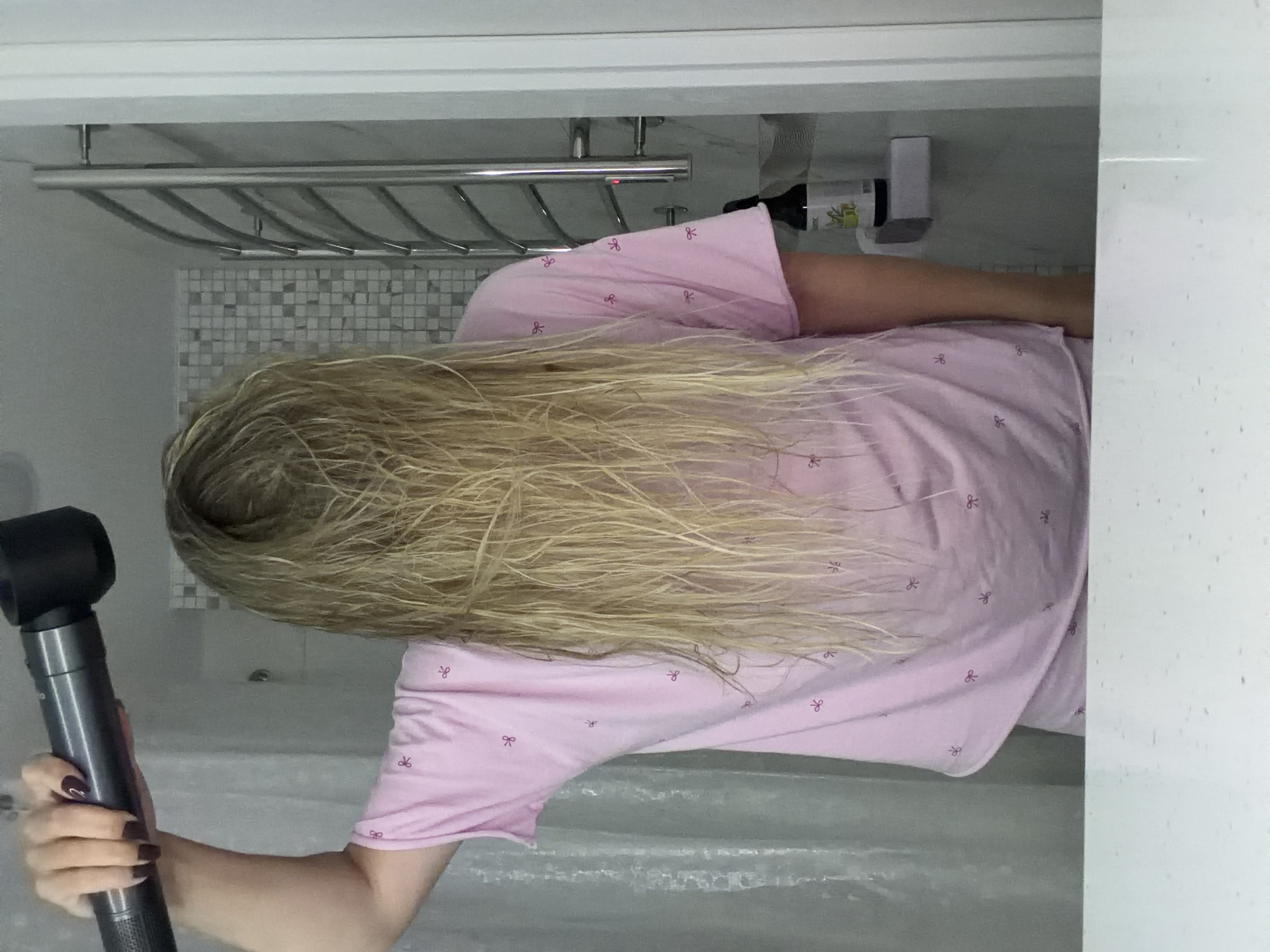 A person with long, wet blonde hair is drying their hair with a dark grey hairdryer. They are wearing a pink t-shirt with a pattern of small red bows. The background shows a tiled wall, a chrome towel rack, a shelf with a dark bottle on it, and a white shower curtain. This scene is not indicative of any specific location in Votkinsk, Russia, or any other city.