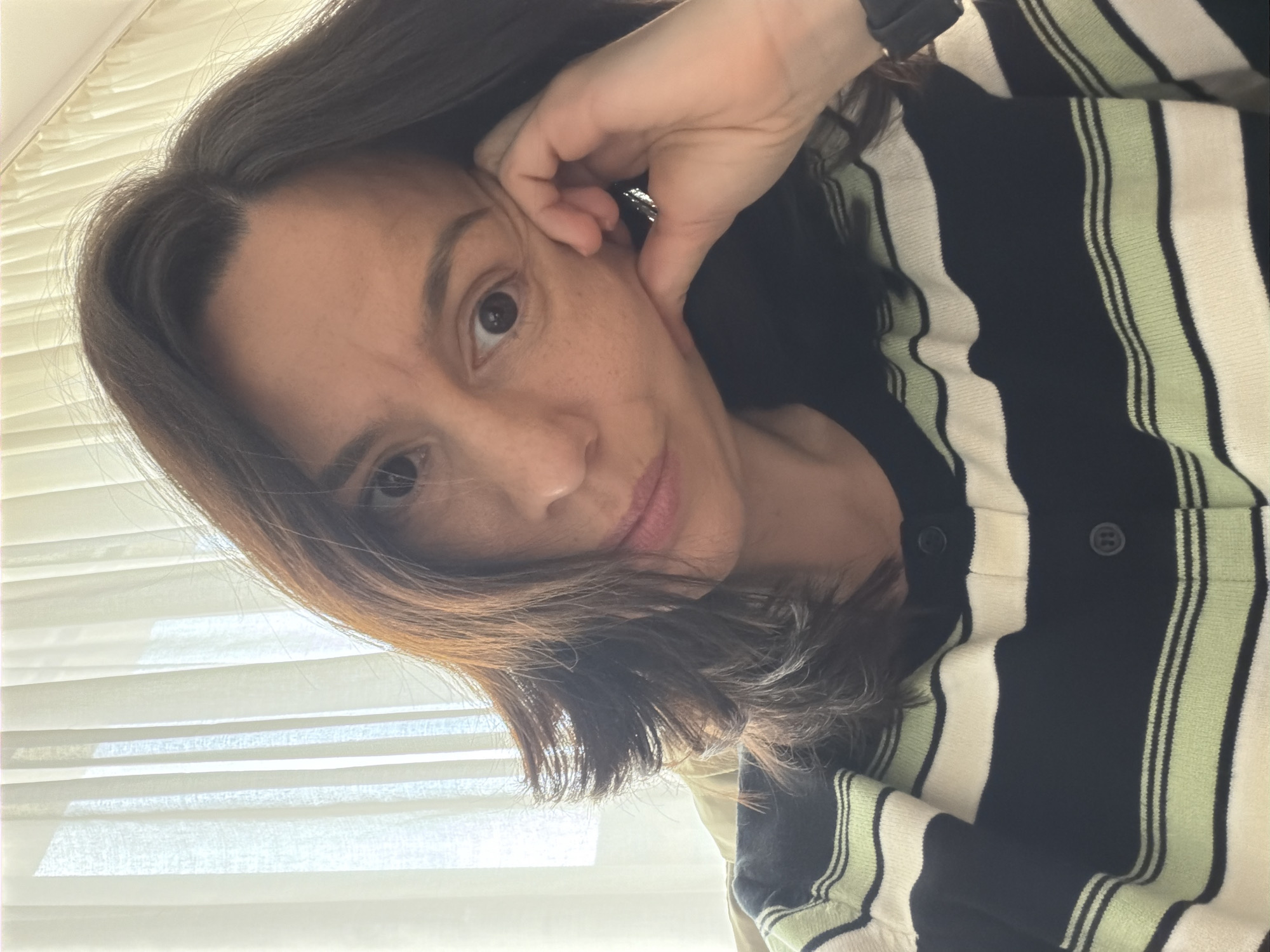 A woman with dark hair is angled towards the camera, resting her head on her hand. She has light skin, brown eyes, and is wearing a striped black, white, and light green cardigan. Behind her is a white, horizontally pleated window covering. The location appears to be indoors. Sofia, Bulgaria is not discernible from the image.