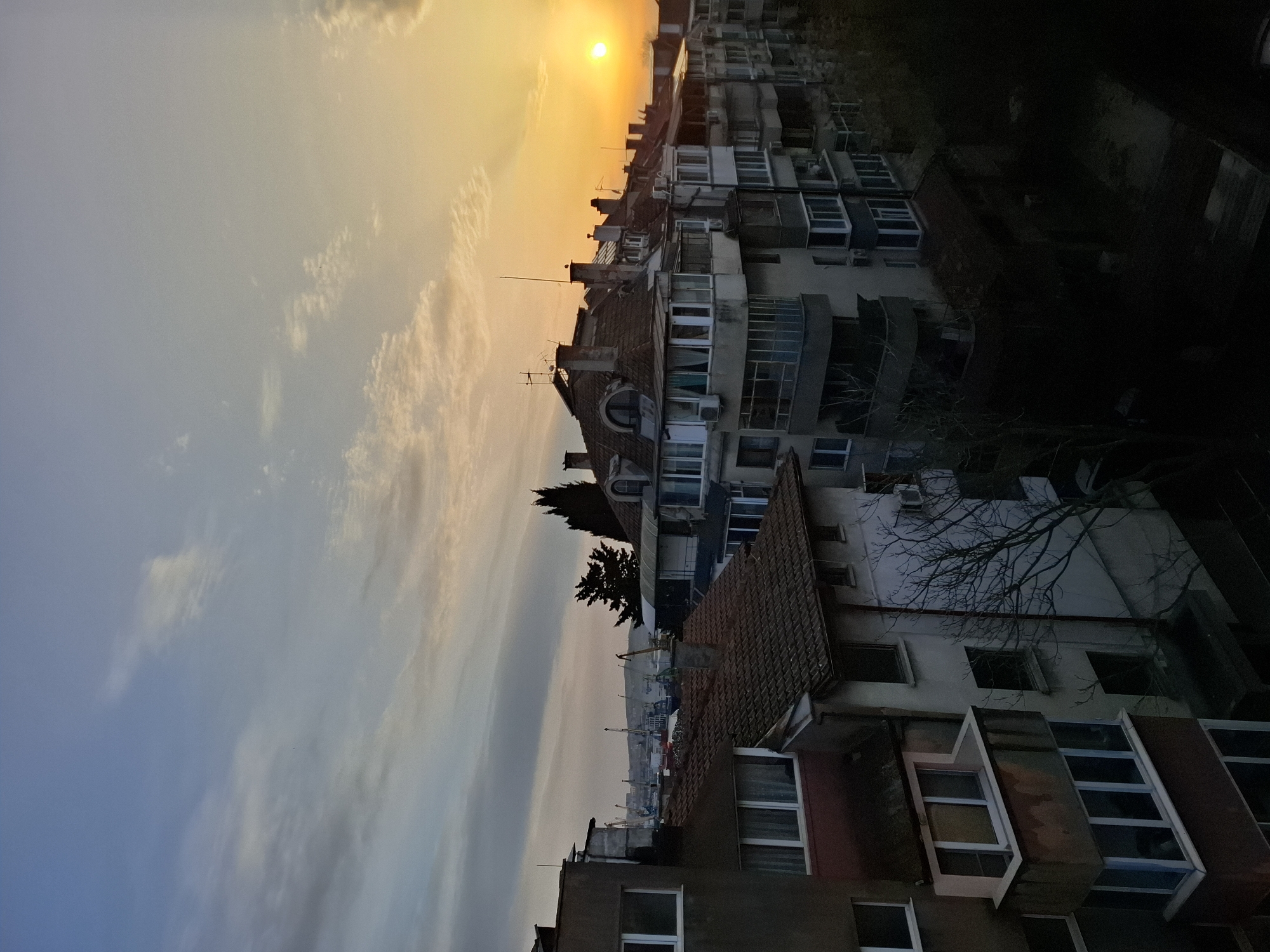 The image depicts a cityscape at sunset, likely in Burgas, Bulgaria, due to architectural styles prevalent in the region. The sun, a bright orb, is setting over a dense cluster of multi-story buildings, casting a warm orange glow on the sky and the upper portions of the buildings. The sky transitions from a light blue with wispy clouds to a yellow-orange hue near the horizon.
The buildings are a mix of residential apartments, constructed from concrete, with variations in design including balconies, windows, and rooflines. Some buildings feature traditional tiled roofs, while others are flat. There are trees interspersed among the buildings, some bare and some evergreen.
In the distance, it is possible to see the silhouette of cranes and possibly a port, implying a location in a coastal city. The lower part of the image fades into shadow, obscuring details of the street level. There are no people visible in the scene.