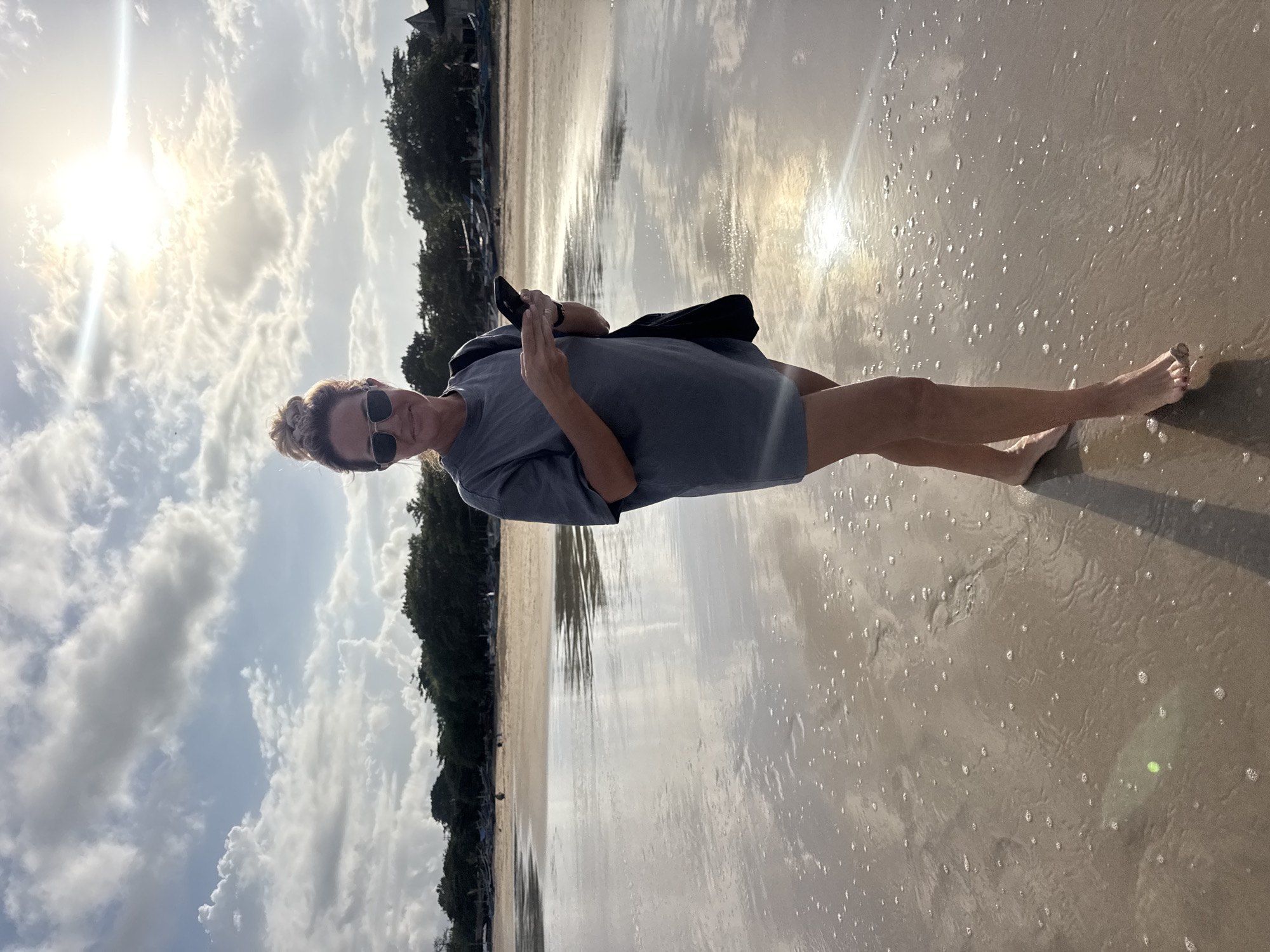 A woman with blonde hair in a bun, wearing sunglasses, a gray t-shirt, and shorts, walks on a beach in Kuta Selatan, Indonesia. She holds a black smartphone in her hands, looking at it as she walks. She is carrying a black bag on her shoulder. The sun is shining brightly in the sky, with scattered clouds. The wet sand reflects the sky and the sun. In the background, trees line the shore, and several boats are visible. The waves are gently lapping at the shore.