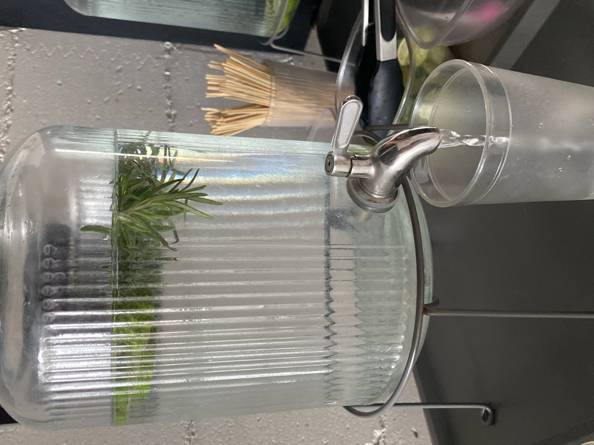The image shows a glass beverage dispenser, likely containing water infused with herbs, being used to fill a clear plastic cup. The dispenser is ribbed horizontally and has a metal spigot. Rosemary is visible inside the dispenser. The dispenser is positioned on a metal stand. In the background, there are wooden skewers in a plastic container, along with some kitchen utensils. The setting appears to be a kitchen or beverage service area. There is no information about North Kuta, Indonesia in the image, so location context cannot be confirmed.