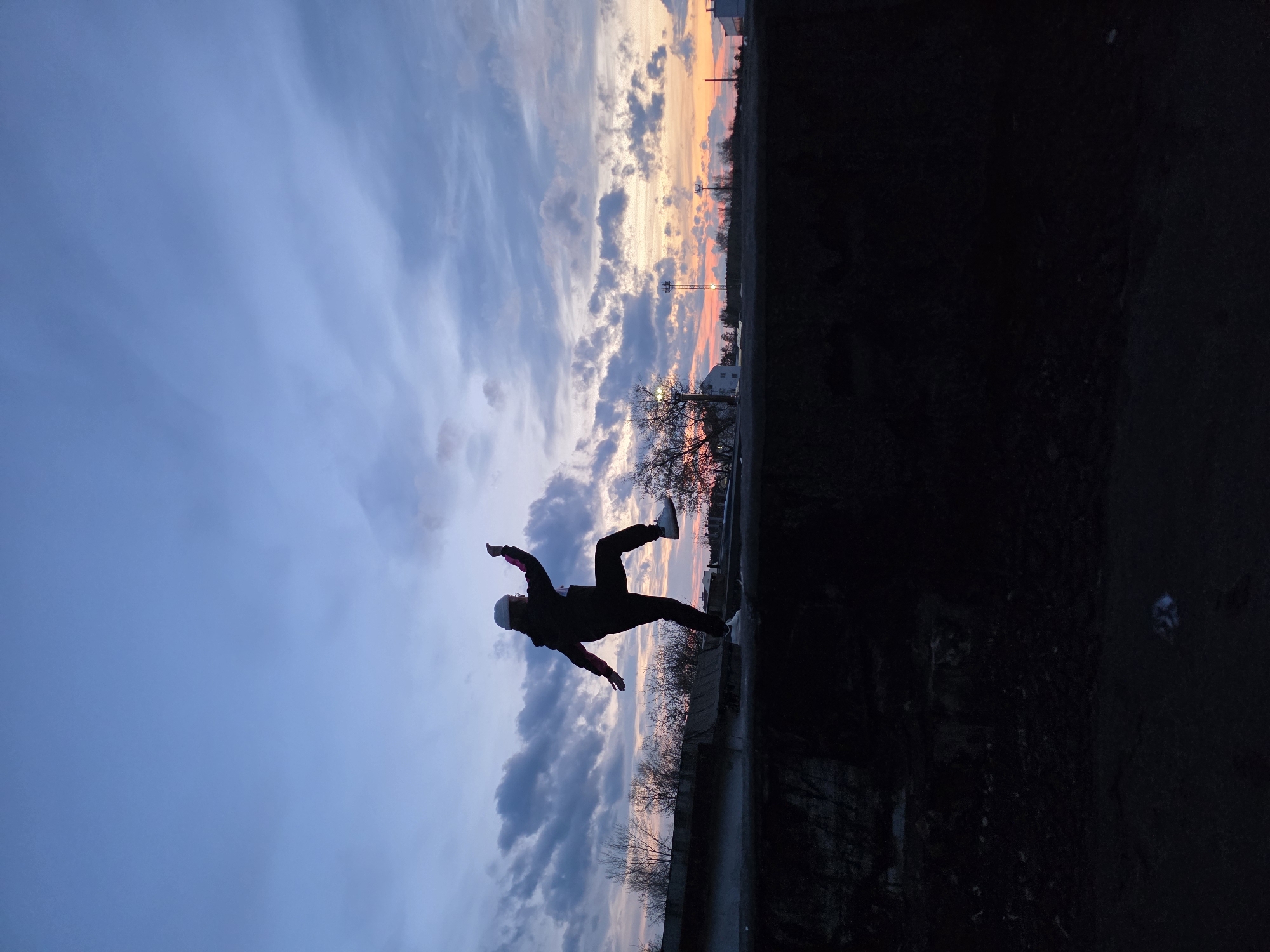 The image features a person silhouetted against a dramatic sunset or sunrise sky in Chaikovsky, Russia. The sky transitions from a soft blue overhead to vibrant orange, pink, and purple hues near the horizon, dotted with varied clouds. The central figure is captured in mid-air, performing a jump or dynamic pose, with arms and legs outstretched. They are wearing dark clothing, a light-colored cap or beanie, and white shoes are visible.
The background shows a distant horizon line with silhouettes of bare trees and some faint structures, possibly buildings or utility poles, against the colorful glow. The foreground and the right side of the image are dark and indistinct, likely representing the ground or a dark wall, creating a strong contrast with the bright sky. The overall scene conveys a sense of movement and freedom against a striking natural backdrop during twilight. No text is visible.