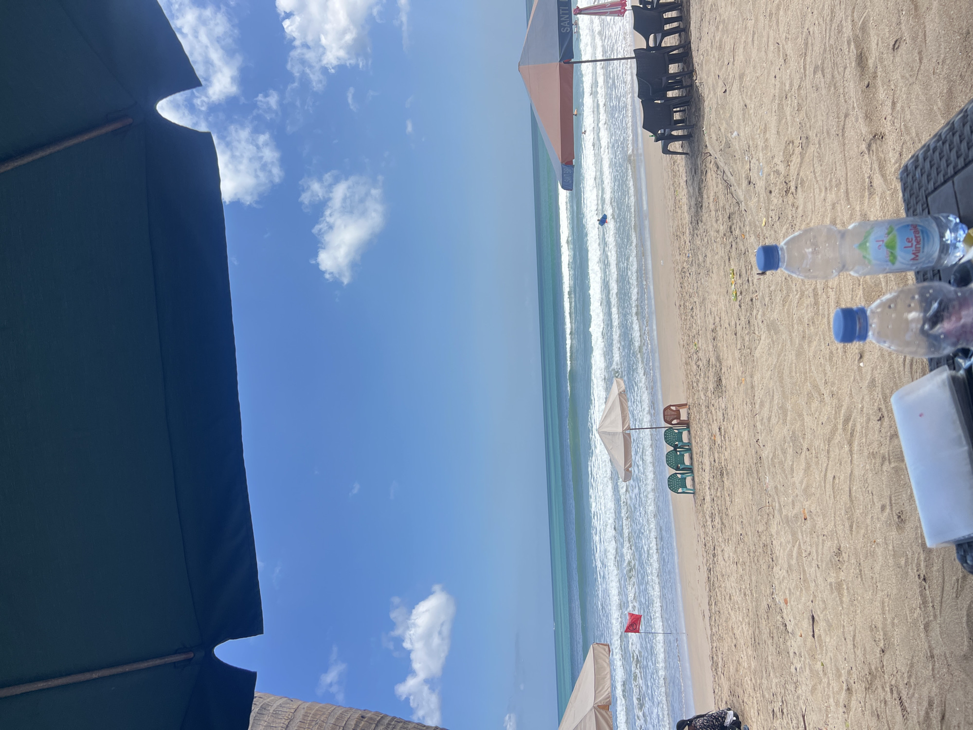 A view of Kuta Beach, Indonesia, from under a dark green umbrella, shows a sandy shore with the ocean in the background. The water is a vibrant turquoise, with waves breaking near the shore. Several beach umbrellas and chairs are set up on the sand. A red flag is planted in the sand near the water's edge, indicating caution. In the foreground, two plastic water bottles sit on a textured surface. The sky is bright blue with scattered white clouds. The scene conveys a sense of relaxation and enjoyment of a tropical beach environment.