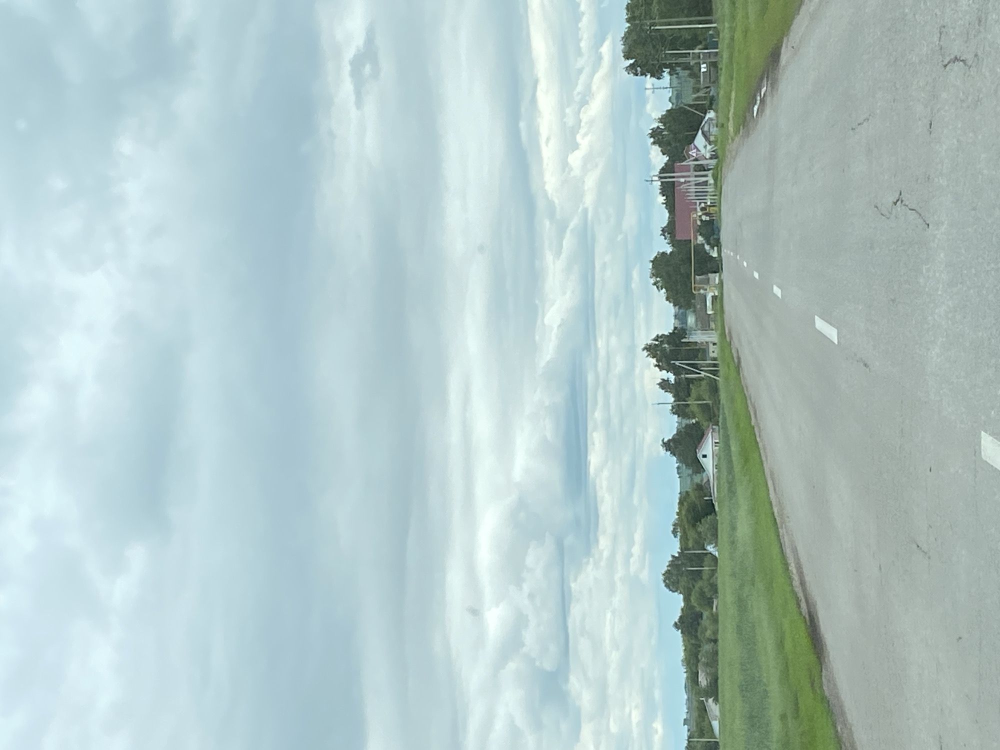 An asphalt road stretches into the distance through a rural landscape under a cloudy sky. The road has white dashed lines marking the lanes. On either side of the road is a vibrant green field. In the middle ground, a cluster of buildings is visible, including houses with red roofs, and trees. The sky is filled with gray and white clouds, with some brighter patches suggesting sunlight breaking through. The scene appears to be in or near the village of Nadezhdino, Russia, as indicated by the environment and architectural style. There are no people visible in the image.