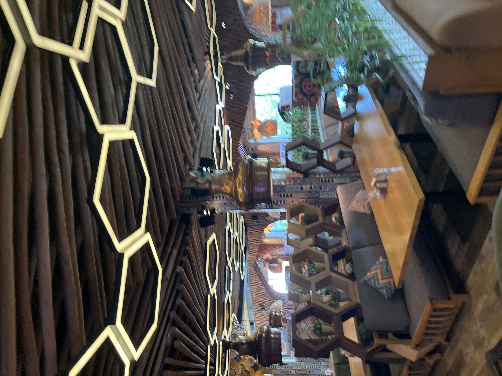 The image shows the interior of a restaurant with a distinctive hexagonal light fixture on a wooden slatted ceiling. The decor features wooden furniture, including booth seating with grey upholstery and a wooden table in the foreground. Hexagonal shelving units with decorative elements and small potted plants are visible. Several ornate brass pendant lamps hang from the ceiling. Through an archway in the background, natural light is visible, suggesting an exit or large windows, with greenery beyond. The overall aesthetic is rustic and modern, with warm wood tones and geometric lighting. The scene is devoid of any people, and no specific action is taking place. The location is identified as a restaurant in Moscow, Russia.