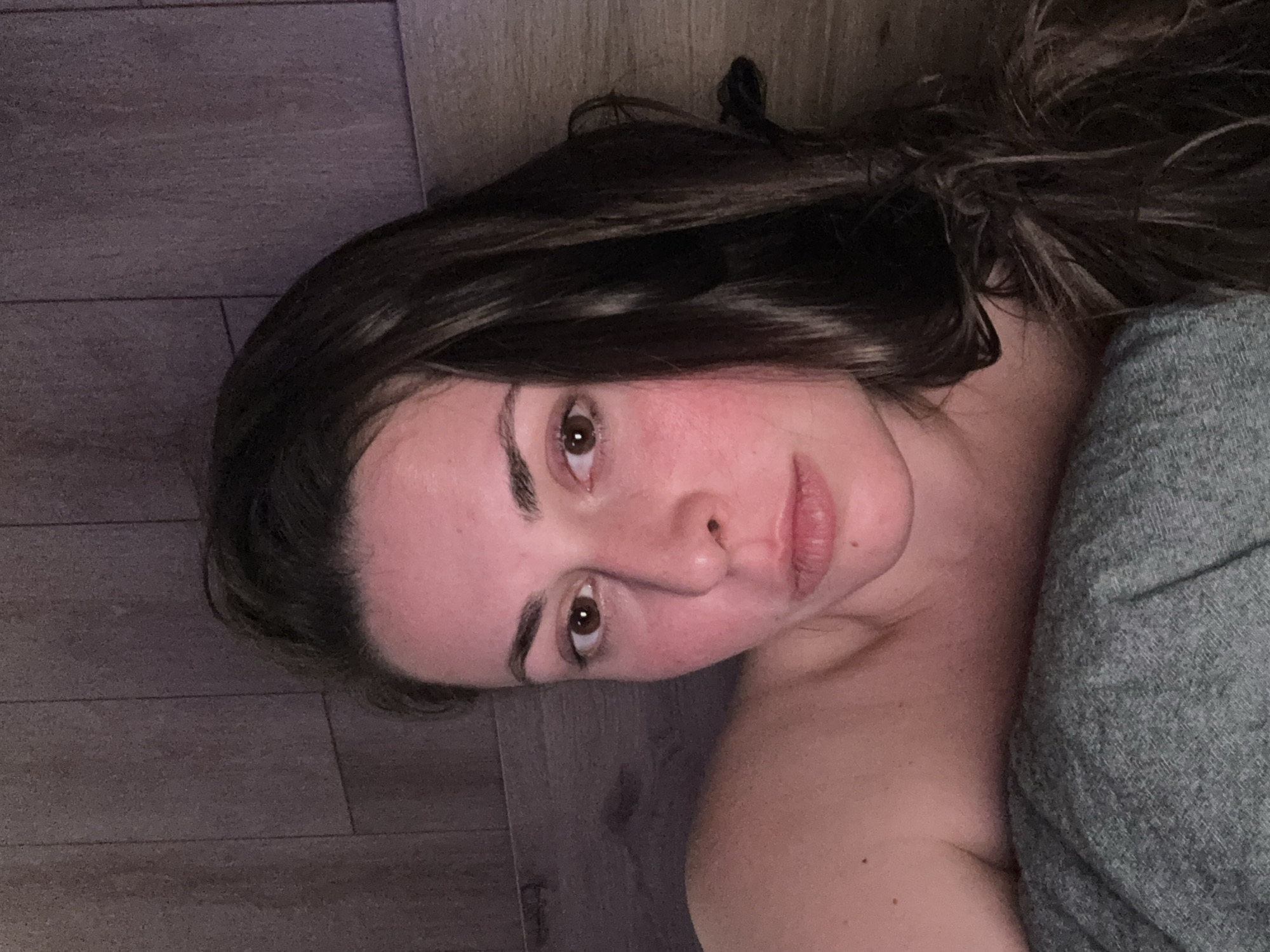 A young woman with dark brown hair is lying down and looking at the camera. She is wearing a grey top and is against a wooden wall. The lighting is dim, and her face is lit from the front.