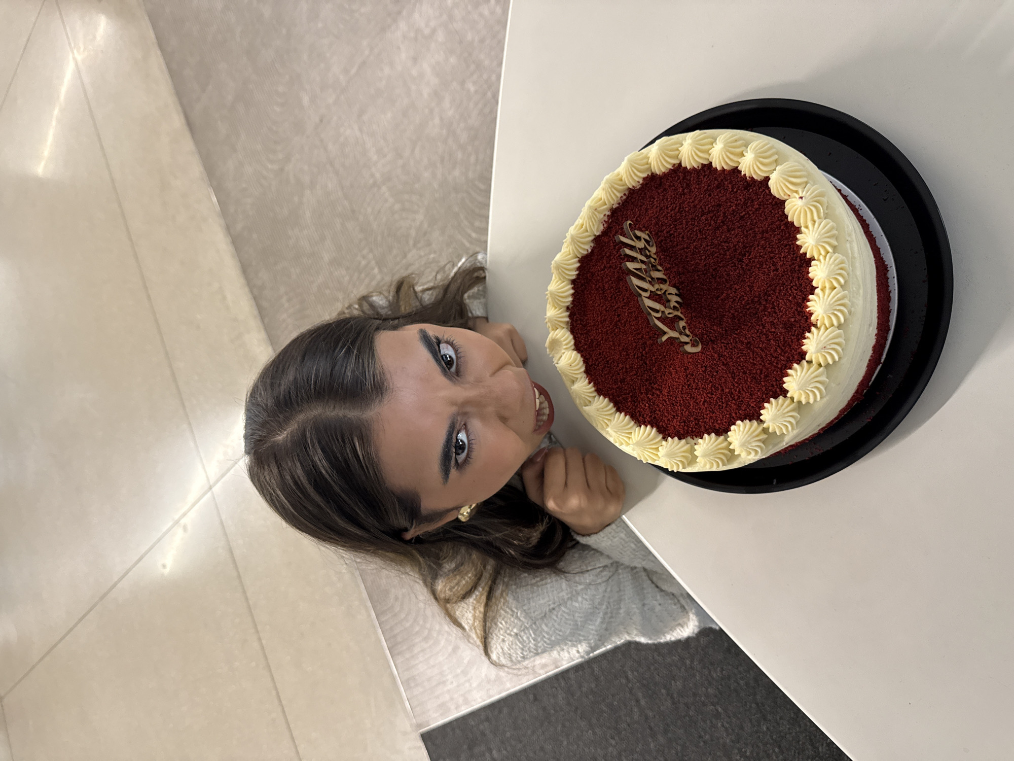 A woman stands next to a white table. On the table, there is a red velvet cake decorated with white frosting and a "Happy Birthday" cake topper. The woman is looking up at the camera, grinning. The location is unknown, but it is in Mexico City, Mexico.