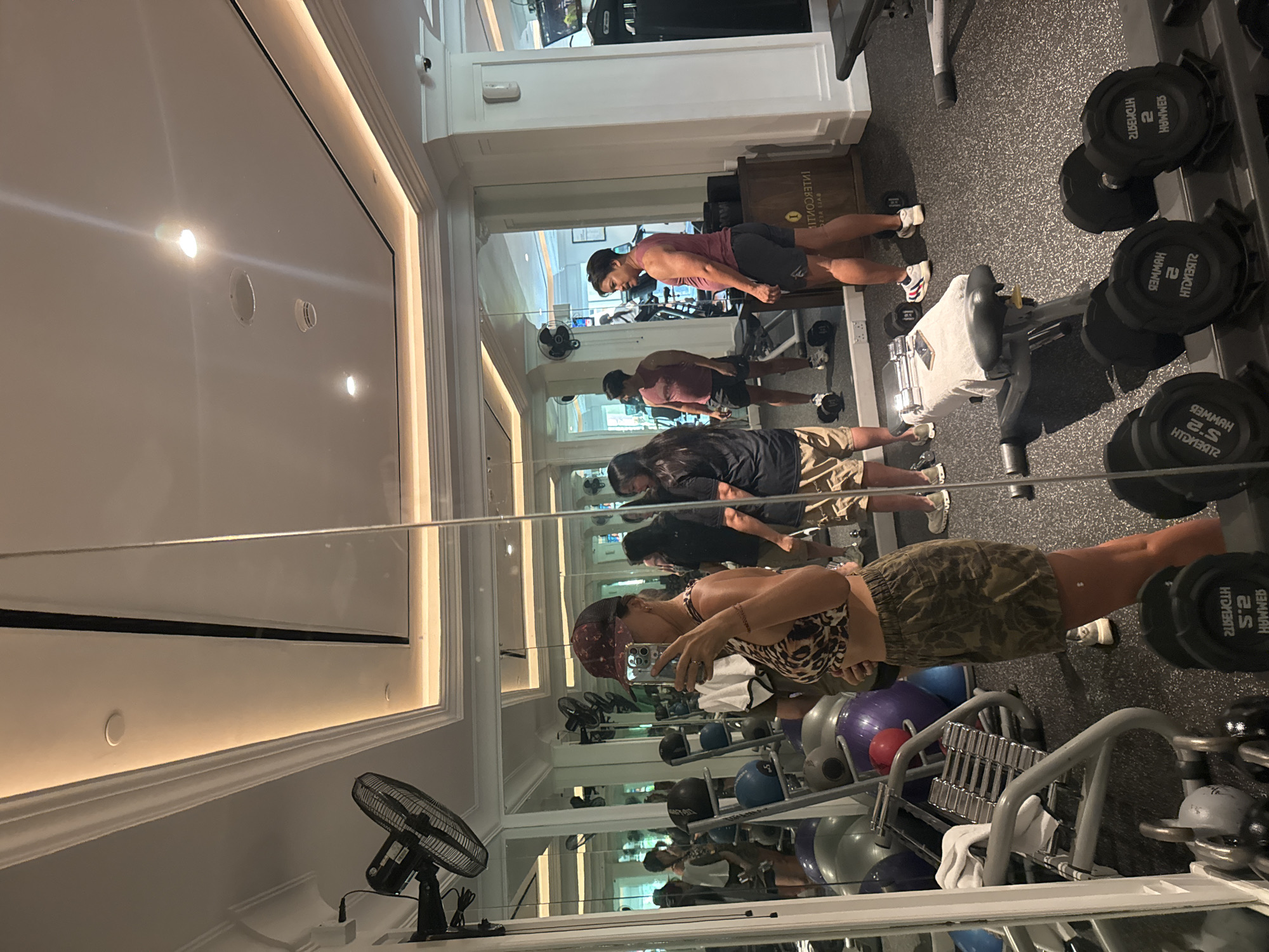 Captured in a well-lit fitness center, the scene showcases a mirrored wall reflecting the gym's interior. Three individuals are engaged in various exercises, with one taking a selfie in the foreground. Weights and exercise balls are visible in the middle ground, and dumbbells are on a rack in the background. The setting is likely a hotel gym or private fitness center, based on its sophisticated layout. The location is suspected to be in Kuta Selatan, Indonesia, given the context.