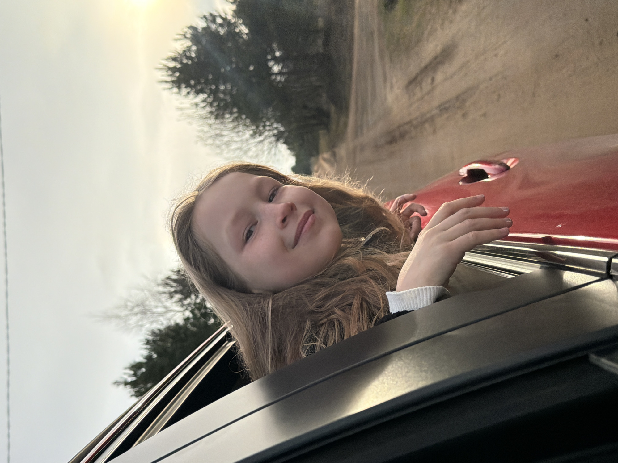 A young girl with long, light brown hair smiles brightly as she leans out of the open window of a red car. Her head is tilted slightly, and her left hand rests gently on the car's window frame. She wears a dark jacket with a white cuff visible at her wrist. Her expression conveys happiness and contentment.
The scene takes place outdoors on what appears to be a dirt or gravel road, suggesting a rural or semi-rural setting, possibly in or around Chaikovsky, Russia. In the background, beyond the car, a line of trees with bare or sparse foliage is visible, with the road extending into the distance. The sky is bright and slightly hazy, indicating daylight, with the sun casting a strong glow from the upper left portion of the image. The bright conditions suggest clear weather. No discernible text is visible in the image.