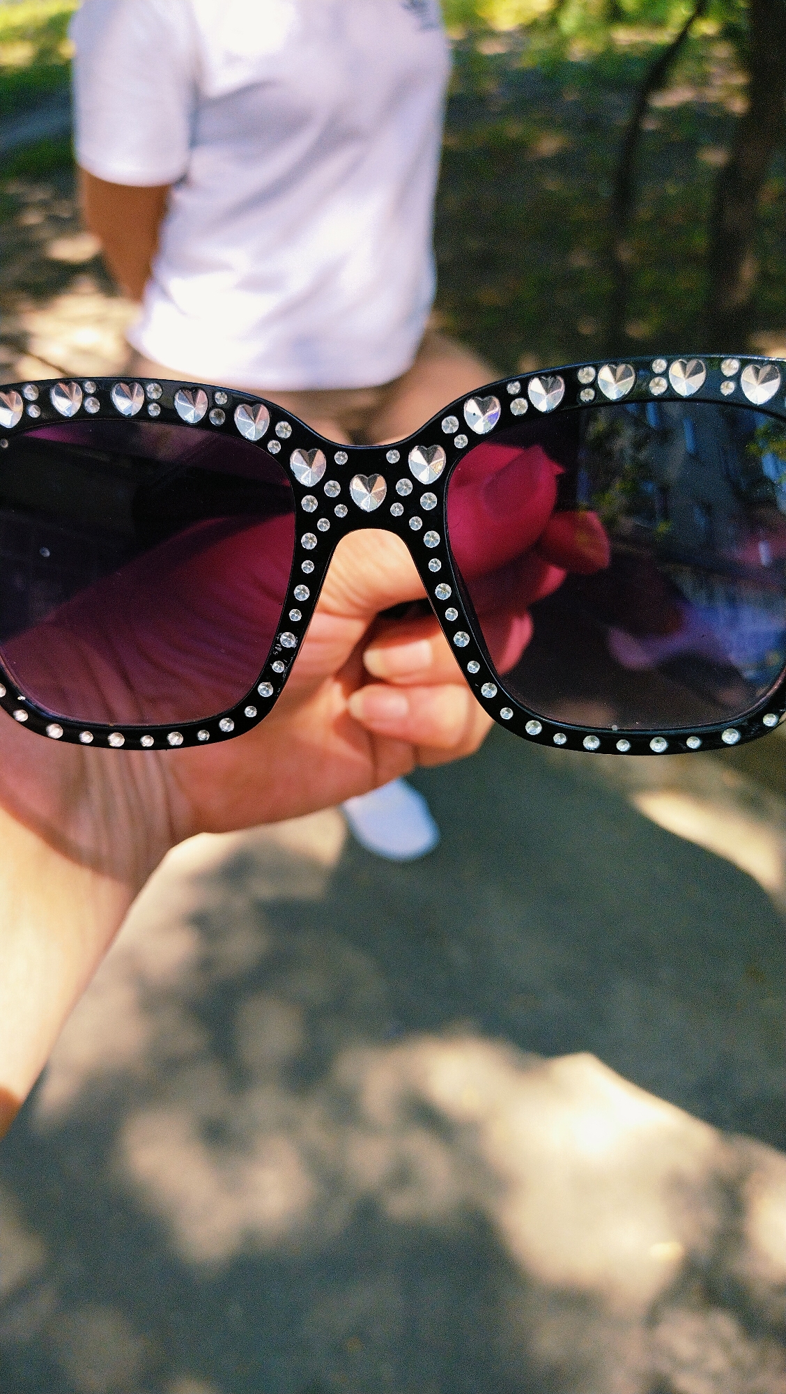 A close-up, slightly angled shot shows a person's left hand holding a pair of black, oversized sunglasses adorned with rhinestones, many of which are shaped like hearts. The sunglasses are held in front of the person's body, obscuring their lower torso and legs. The person is wearing a white t-shirt and appears to be sitting outdoors, with dappled sunlight and shadows from trees falling on the ground and the subject. The background is out of focus, with hints of green foliage and a blurry view of a paved surface. The image appears to be taken in the city of Horishni Plavni, Ukraine, suggested by the outdoor environment with trees and a paved area, common in parks or public spaces within the city. The overall impression is casual and summery.
