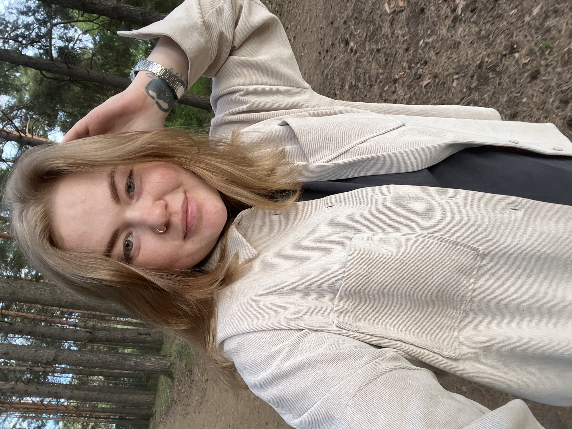 A young fair-skinned person with light brown hair is posing for a selfie in an outdoor wooded area. The individual is wearing a light beige corduroy-like jacket over a dark-colored shirt. The jacket is buttoned, and a pocket is visible on the left side. The person is wearing a watch with a silver band on their left wrist and a tattoo just below the watch. The left hand is partially raised and touching the person's head. A small nose ring is visible. The background shows tall trees indicative of a forest. Based on the provided information, the location is likely near Chaikovsky, Russia. The ground appears to be covered with leaves and dirt. The image has been rotated counter-clockwise.