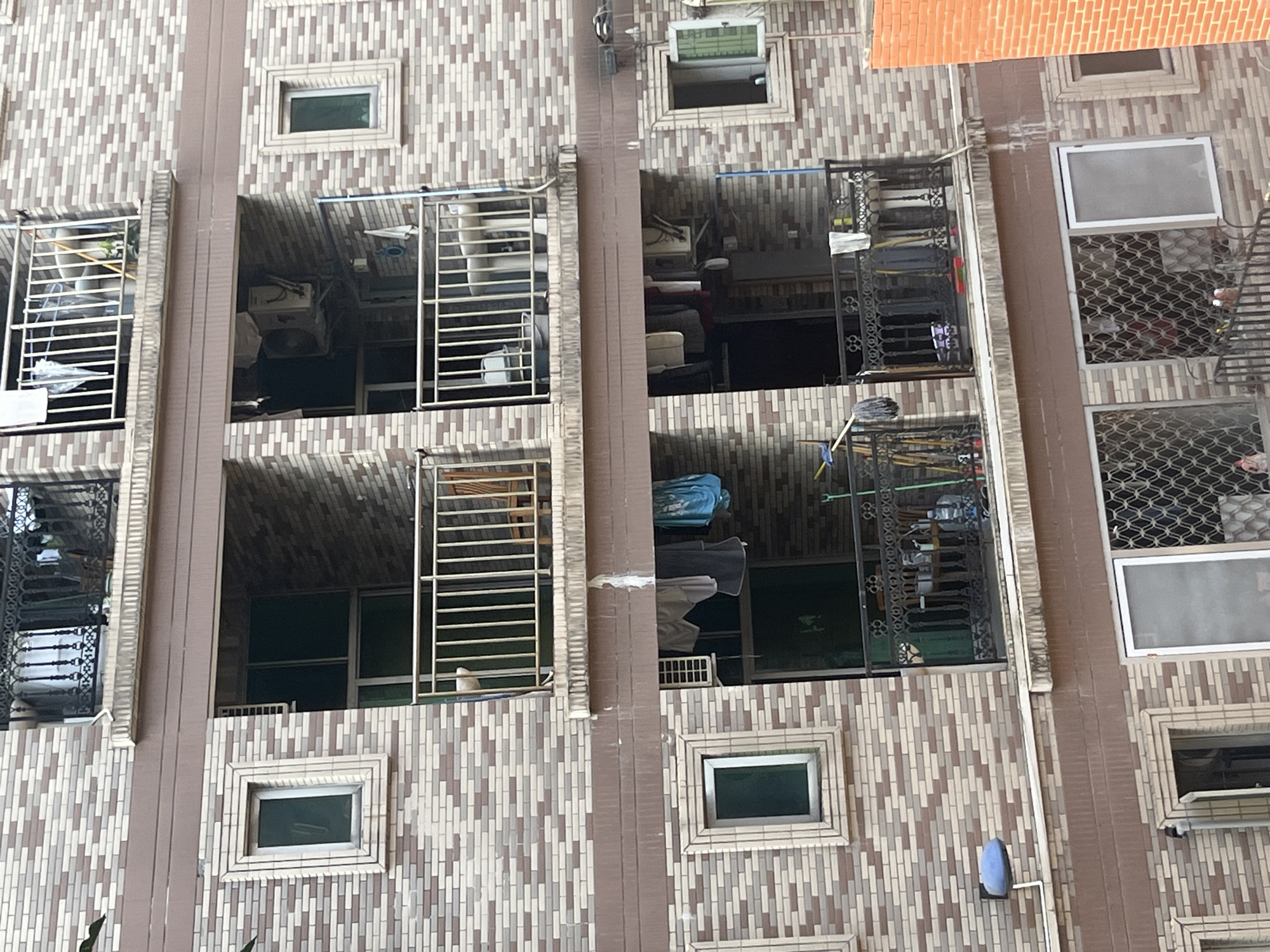 The image depicts the exterior of a multi-story apartment building with a brick facade, located in Yuzhong District, China. The building features several balconies, some with railings and others enclosed. Laundry is visible hanging on clotheslines on some of the balconies. Air conditioning units are mounted on the exterior walls. Windows of various sizes are present, some open and some closed, and some are covered with decorative metal grates. The building's architecture suggests a residential area. The lighting conditions indicate it is daytime. No people are directly visible in the frame.