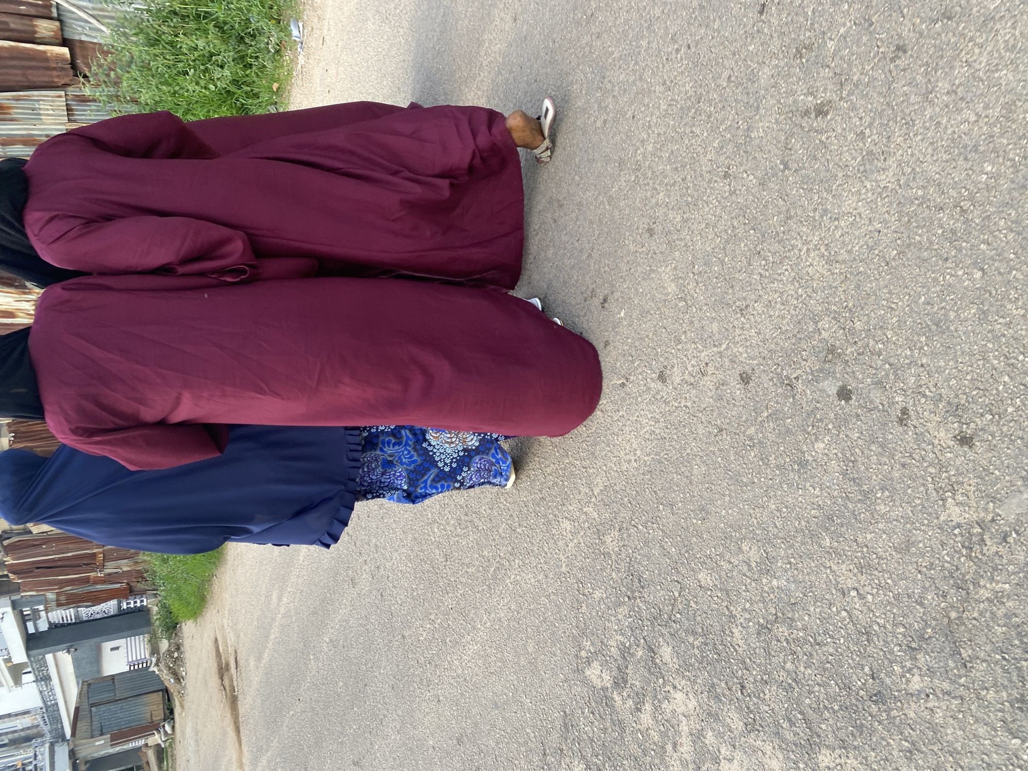In the city of Jos, Nigeria, three women are walking on a paved road. The woman on the left is wearing a navy blue hijab and a patterned blue skirt. The other two women are wearing matching burgundy abayas. One of the women in the abaya is wearing sandals. The background shows a building with corrugated metal siding and some greenery.