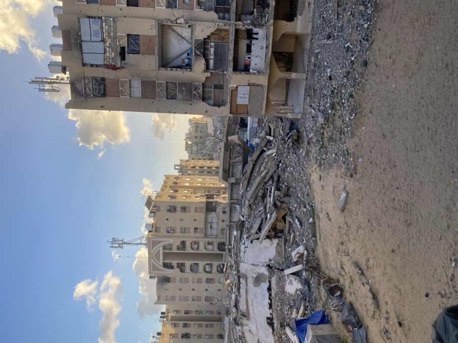 The image depicts a scene of extensive urban destruction in Khan Yunis, Palestinian Territory. In the foreground, a sandy, barren ground transitions to a midground dominated by large piles of concrete rubble, twisted rebar, and shattered building materials, indicating the complete collapse of at least one structure. To the right of the frame, a multi-story, tan-colored residential building stands partially destroyed, its facade perforated with numerous holes and large sections of walls missing, exposing interior rooms. Windows are either blown out, boarded with various materials, or covered with makeshift tarps. Clothes are visible hanging to dry within an exposed lower-level apartment. In the background, several similar multi-story buildings, also appearing tan or beige, exhibit significant damage including impact holes, missing windows, and damaged balconies. Some buildings feature decorative arched elements near their rooftops, which are also topped with various antennas and communication equipment. The sky above is clear blue with scattered white clouds, illuminated by a warm, golden sunlight consistent with either early morning or late afternoon. No individuals or active interactions are visible in the scene.