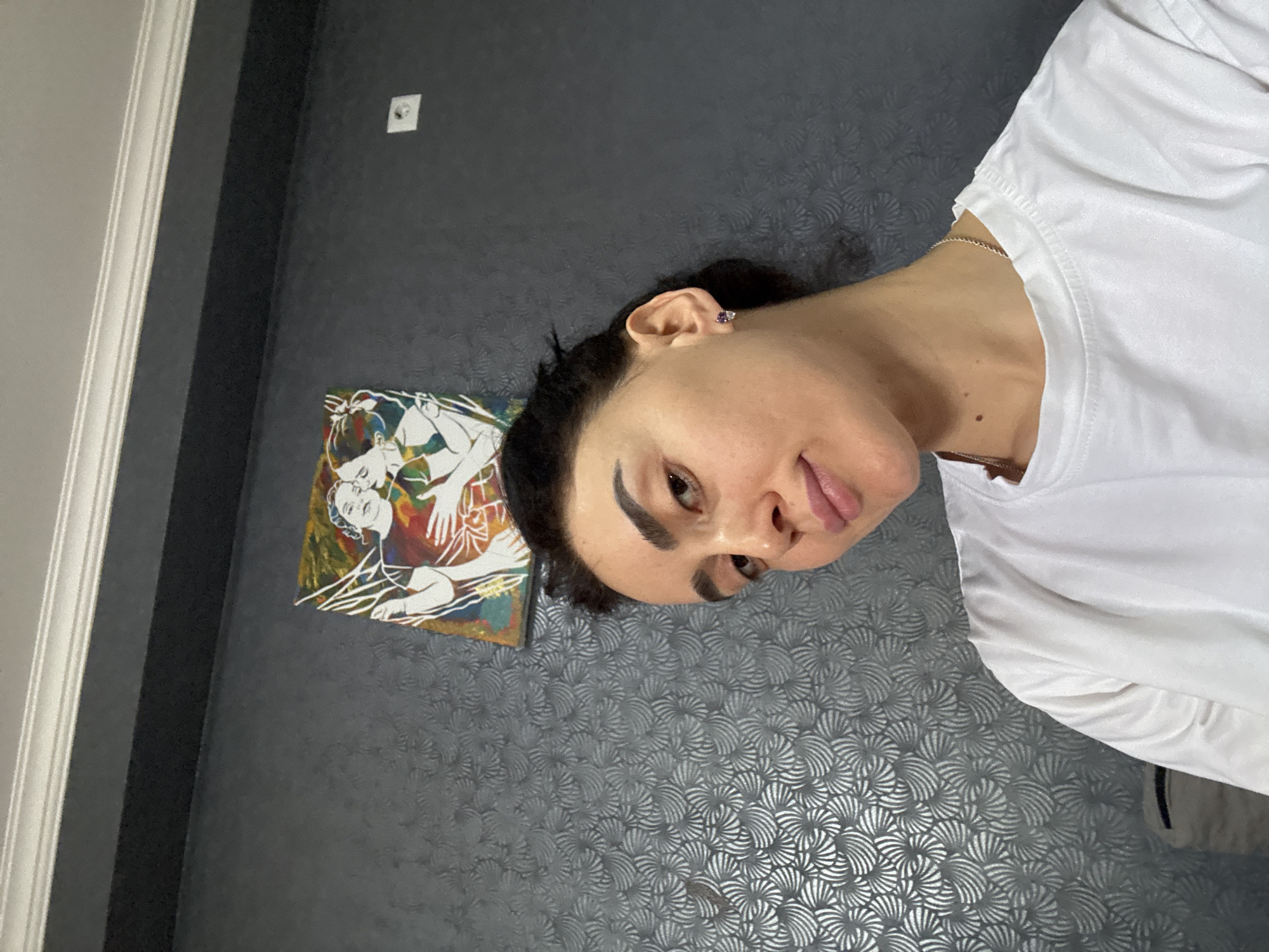 A person with dark hair and a white t-shirt is indoors against a gray wall with a patterned texture. The person is wearing an earring and a necklace. A painting hangs on the wall to the left of the person. The setting appears to be a room, possibly in a residential building. There is no information that indicates the location in Tbilisi, Georgia.