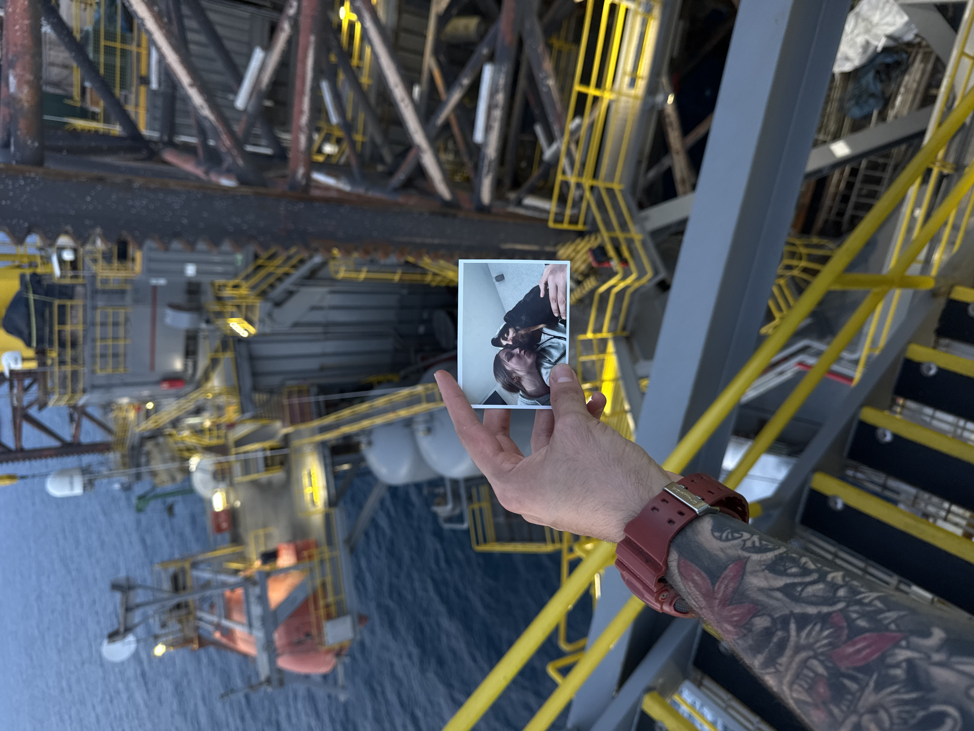 A person's left arm, adorned with a red watch and an elaborate tattoo, is in the foreground of the image, holding a small photograph. The photograph displays a close-up of a woman and a dog, both appearing to be indoors. The background of the main image is a large, industrial offshore oil rig situated above a body of dark blue water. The oil rig consists of a complex structure of metal beams, platforms, yellow railings, and various pieces of equipment, including a bright orange lifeboat. The scene is captured from an elevated perspective, looking down onto the rig and the water. The lighting suggests it may be dusk or early morning. The location is not specified within the city of Unknown, Italy.