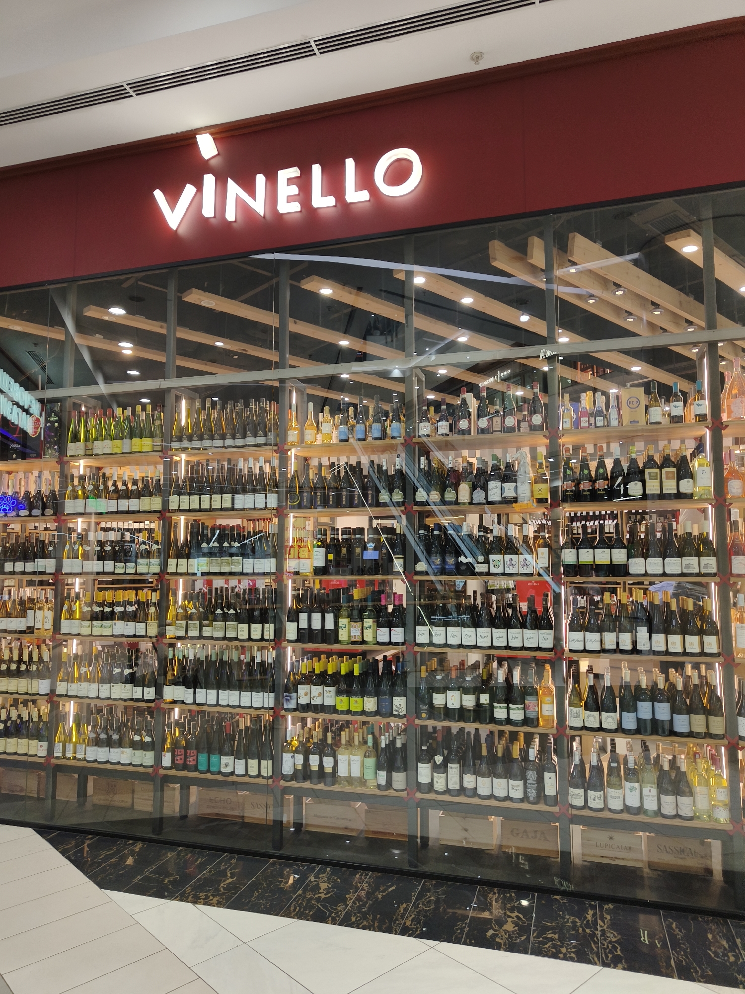 The image shows the interior of a wine shop called "VINELLO" in Sofia, Bulgaria. The shop features a large, well-stocked display of wine bottles arranged on multiple shelves behind a glass storefront. The lighting is bright and highlights the rows of wine bottles, which appear to be a mix of red, white, and rosé wines, as well as possibly other spirits.
The storefront is made of large glass panels, allowing a clear view of the merchandise inside. The shelves are organized with wooden accents, and the bottles are neatly arranged, creating an appealing display. Some of the bottles are in wooden crates at the bottom of the display.
The shop's name, "VINELLO," is prominently displayed above the storefront in large, illuminated white letters against a dark red background. The interior ceiling features a modern design with exposed wooden beams and track lighting. The floor in front of the store is a mix of white tiles and black marble with a subtle pattern.
There are no people visible in the image, suggesting the photo was taken during business hours when the shop is open, or perhaps just before opening or after closing. The overall impression is one of a modern and well-curated wine retail environment. The time of day is not explicitly indicated, but the indoor lighting suggests it could be any time. The setting is clearly a commercial space within a building, likely a shopping mall or a street-level retail establishment.
