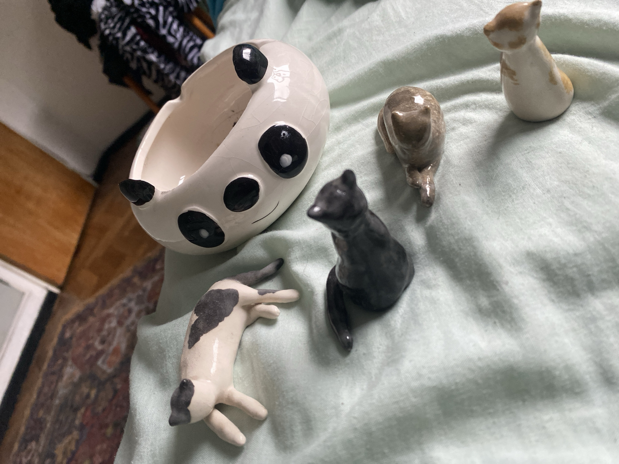 The image shows a collection of small ceramic figurines on a light green fabric surface. There is a panda-shaped bowl and four cat figurines of different colors. The cat figurines are a tan-and-white cat standing, a tan cat lying down, a black cat sitting, and a white-and-black cat lying down. The background includes a wooden floor, a rug, and a striped piece of furniture. The image does not provide any information about the city of Buenos Aires, Argentina.