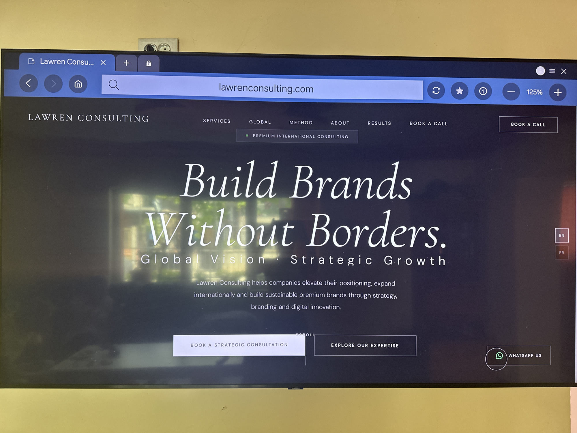 A television screen displays a website for "Lawren Consulting." The website features the tagline "Build Brands Without Borders" and offers services related to global vision and strategic growth. Navigation elements like "Services," "Global," "Method," "About," and "Results" are visible, along with a "Book a Call" button. The scene is captured in a room with a yellow wall and a power outlet above the television. The television itself is a Samsung model.