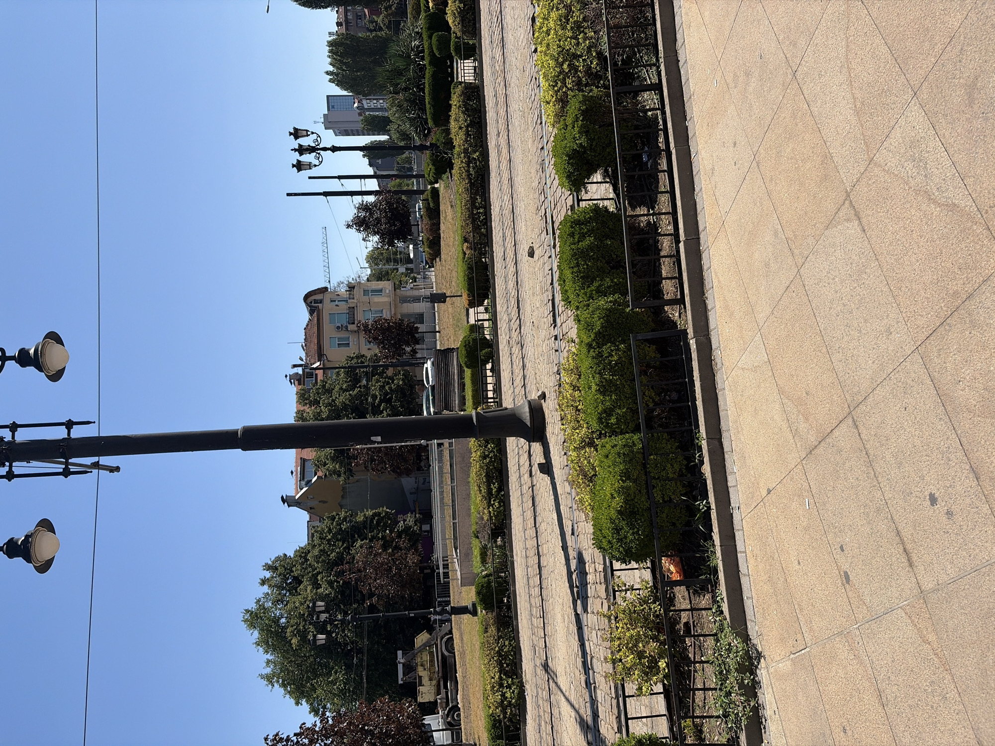 The image depicts a street scene in Sofia, Bulgaria, specifically on the tram tracks near the National Palace of Culture (NDK). The foreground is a paved area with large, square, light-colored tiles. Beyond this, a black metal fence separates the paved area from a strip of green grass and well-maintained hedges. Tram tracks are visible, laid in a cobblestone surface, stretching across the mid-ground of the image. A large, dark lamppost with multiple lights stands prominently in the center of the frame, casting a shadow on the tracks. In the background, trees with lush green foliage line the street. Several buildings are visible, including a multi-story building with a terracotta-colored roof and balconies, and taller modern buildings further in the distance. A yellow construction vehicle is parked on the grass area to the left. The sky is clear and bright blue, indicating a sunny day. There are no people visible in the image.