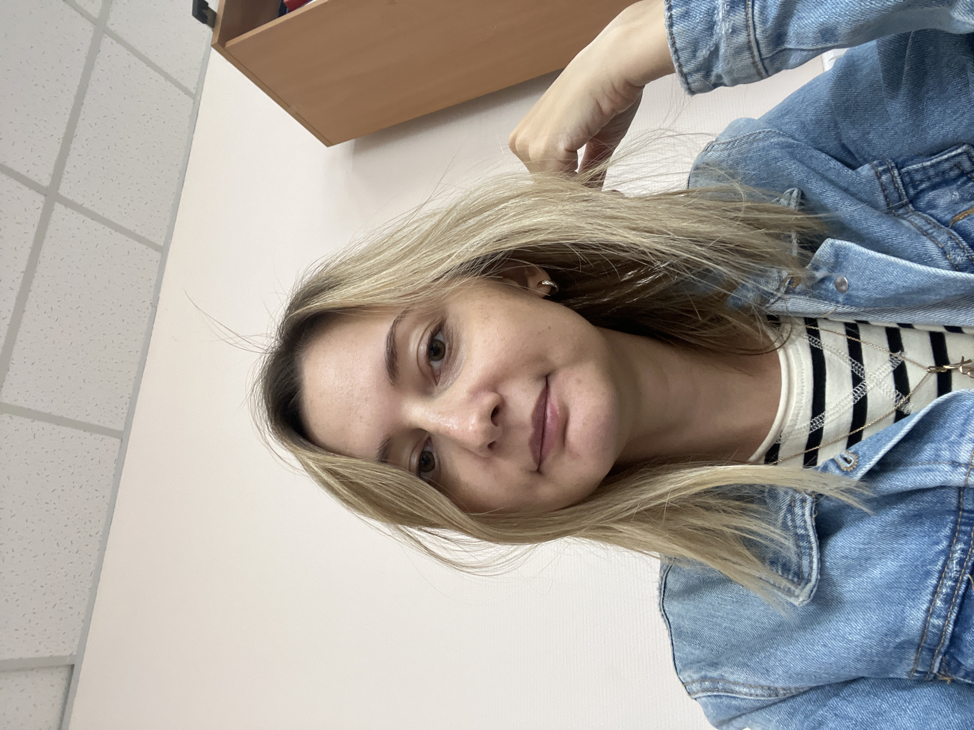 A woman with blonde hair is sitting at a desk in a room. She is wearing a denim jacket over a striped shirt and a necklace. The room has a white wall and a drop ceiling with fluorescent lighting. There is a wooden shelf unit to the right of the woman. The image appears to be a selfie taken indoors. There is no information to indicate the location within the city of Volgograd, Russia.