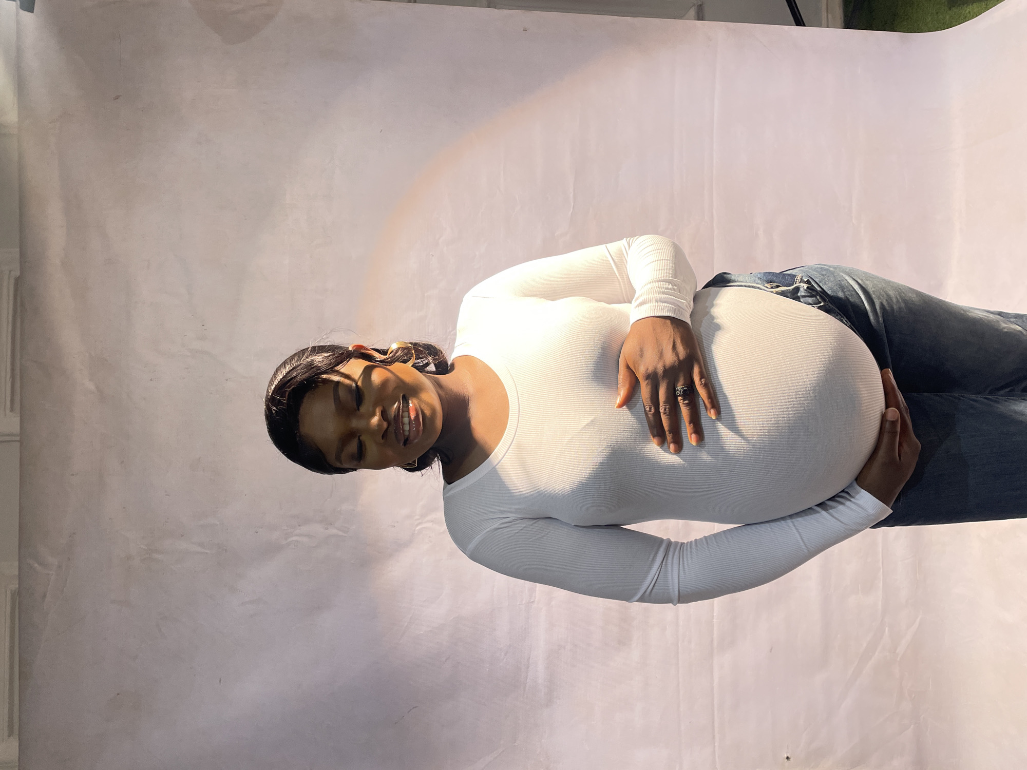 A pregnant woman lies in repose against a white backdrop. The woman wears a white long-sleeved shirt and jeans, her hands gently placed on her pregnant abdomen. Her eyes are closed, and she has a slight smile. The location is a photo studio or similar space with a plain white background. The location is Jos, Nigeria.