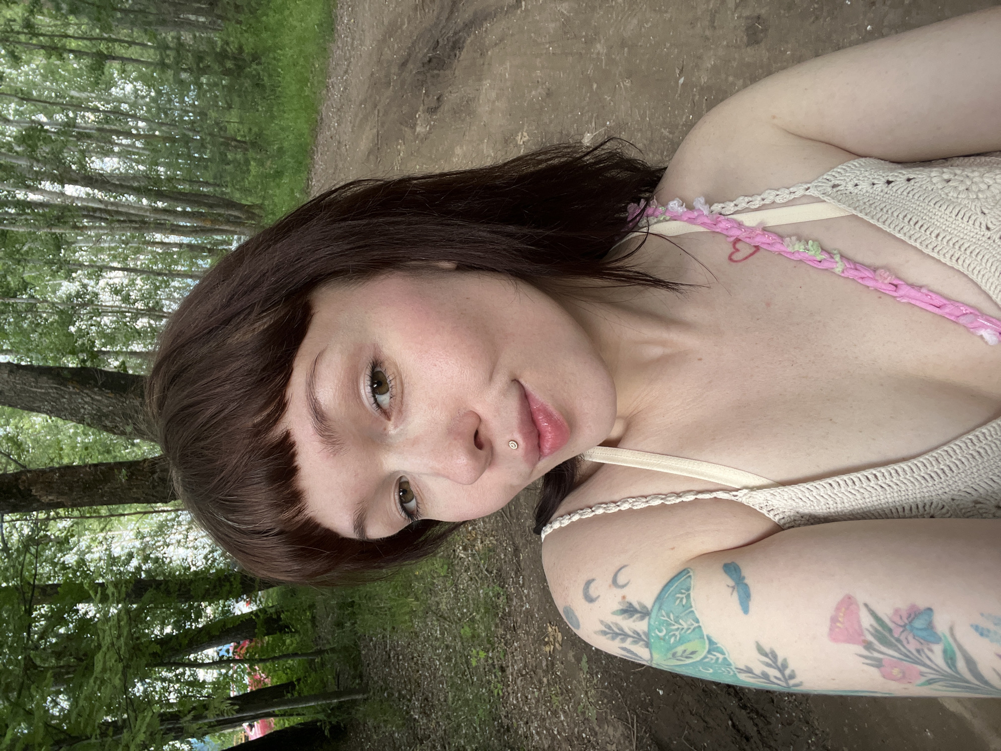 An outdoor, medium close-up selfie shows a young woman with dark brown hair and bangs, a nose ring, and tattoos on her left arm. She is wearing a cream-colored, crocheted top with thin straps and a pink braided embellishment. The background is a dirt road surrounded by trees and greenery, suggesting a natural or park-like setting. The lighting indicates it is daytime. The image does not contain any information that allows for a specific location within the city of Chaikovsky, Russia, to be identified.