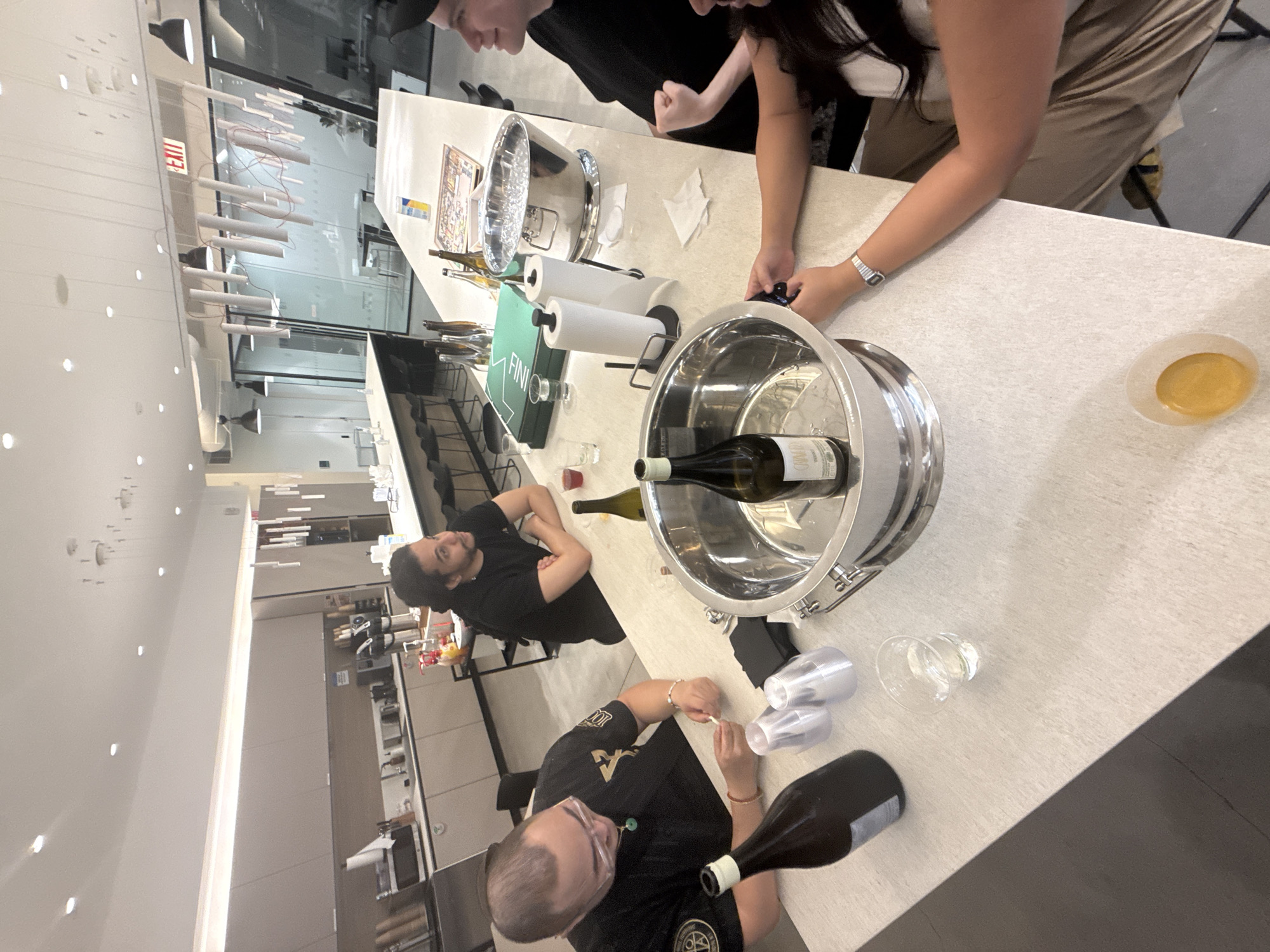 An indoor scene captures four individuals gathered around a light-colored kitchen island or bar, suggestive of a modern office kitchen or event space. The setting is bright, illuminated by overhead recessed lights and possibly natural light through glass partitions.
The central activity appears to be a casual social gathering or wine tasting. Two people stand behind the counter: one partially visible on the right, wearing light pants and a dark top, and another in a dark top holding a cell phone. On the opposite side of the counter, two men are seated or leaning. One has dark hair and a beard, wearing a dark shirt. The other, in the foreground, wears glasses, a dark shirt with an emblem, and appears to be arranging clear plastic cups.
Prominently displayed on the counter are two large metallic wine buckets, one holding a white wine bottle with "CABERNET SAUVIGNON" visible on its label, and several other wine bottles. Other objects include a paper towel holder with white rolls, a small clear glass, a red cup, a dish containing a yellow liquid, and a paper menu or board with "FINI" written on it.
In the background, a modern interior features glass walls, minimalist lighting fixtures, and a bright white ceiling. A red "EXIT" sign is clearly visible above a doorway beyond the counter. Further back, kitchen cabinetry and appliances can be seen. The atmosphere appears relaxed and social. The bright lighting suggests it is daytime.